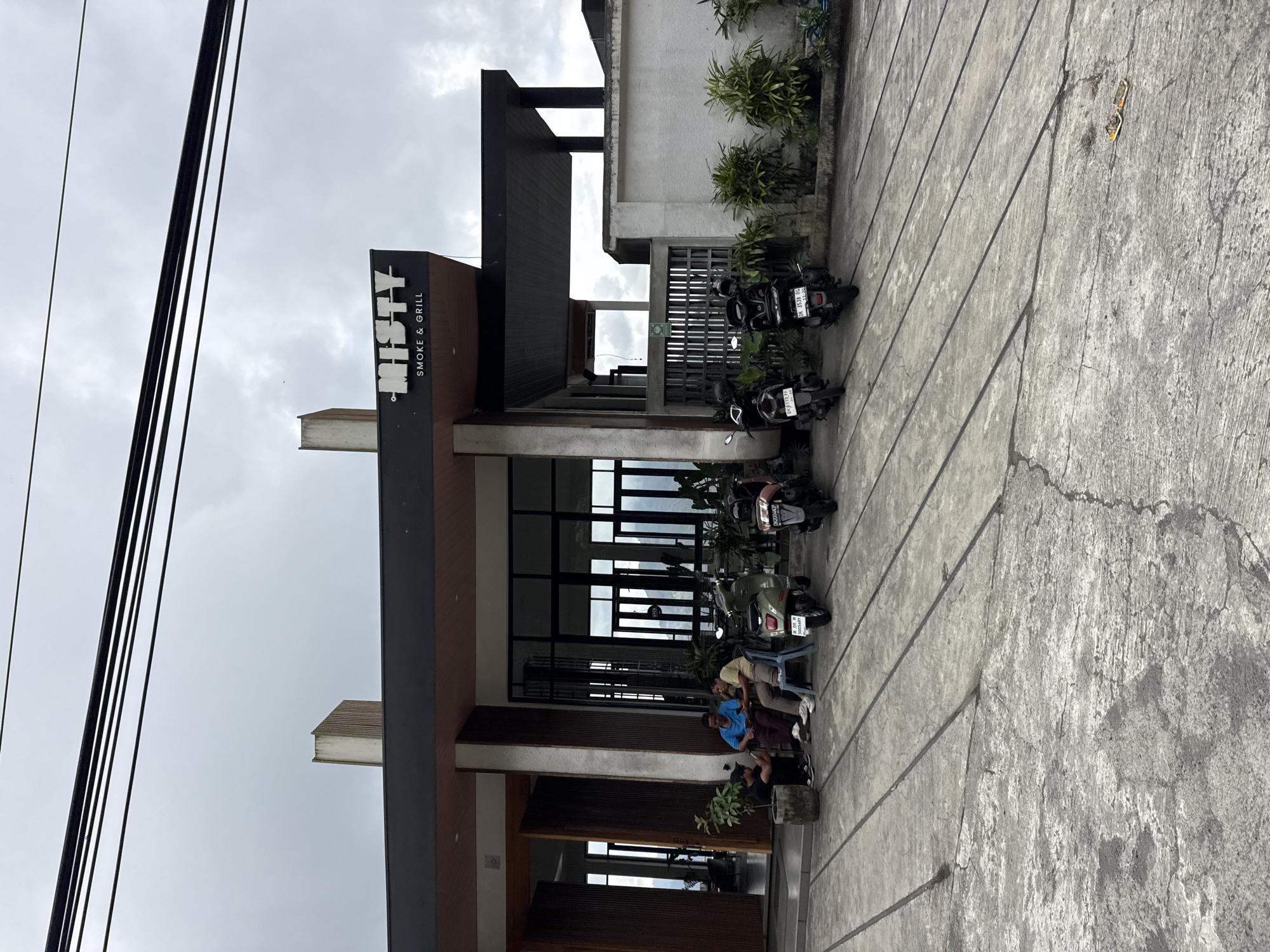 The image shows the exterior of a restaurant called "MISTY SMOKE & GRILL" in Kintamani, Indonesia. The restaurant has a modern design with a dark-colored facade and large glass windows. Several motorcycles are parked in front of the restaurant, suggesting it's a popular spot.
In the foreground, a group of people are seated on a bench outside the restaurant. Their posture and casual attire suggest they are either waiting for a table, taking a break, or socializing. The atmosphere appears relaxed and informal.
The time of day is likely daytime, given the ambient light, but the sky is overcast, indicating a cloudy or possibly rainy weather condition. The concrete pavement in front of the restaurant is marked with diagonal lines, possibly for drainage or decorative purposes. Lush green plants are visible around the building, adding a touch of nature to the urban setting.
The sign for "MISTY SMOKE & GRILL" is prominently displayed, identifying the establishment. The text "SMOKE & GRILL" suggests the type of cuisine offered. The overall scene captures a typical day at a restaurant in a potentially tropical location.