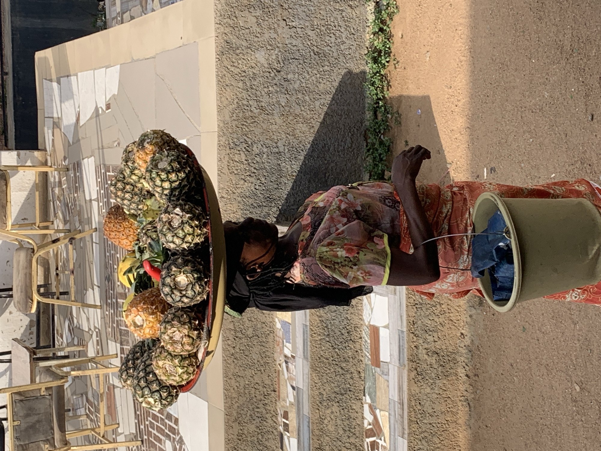 An elderly, dark-skinned woman is standing on a dirt path, likely near New Stadium, Nigeria. She is wearing a floral-patterned dress and a black scarf, with glasses on. A large round tray balanced on her head is loaded with pineapples and some bananas. Beside her, a large green bucket contains what appears to be folded denim clothing. The background includes a textured beige wall, a partial view of a building with light-colored, mosaic-like details, metal chairs, and some sparse greenery.