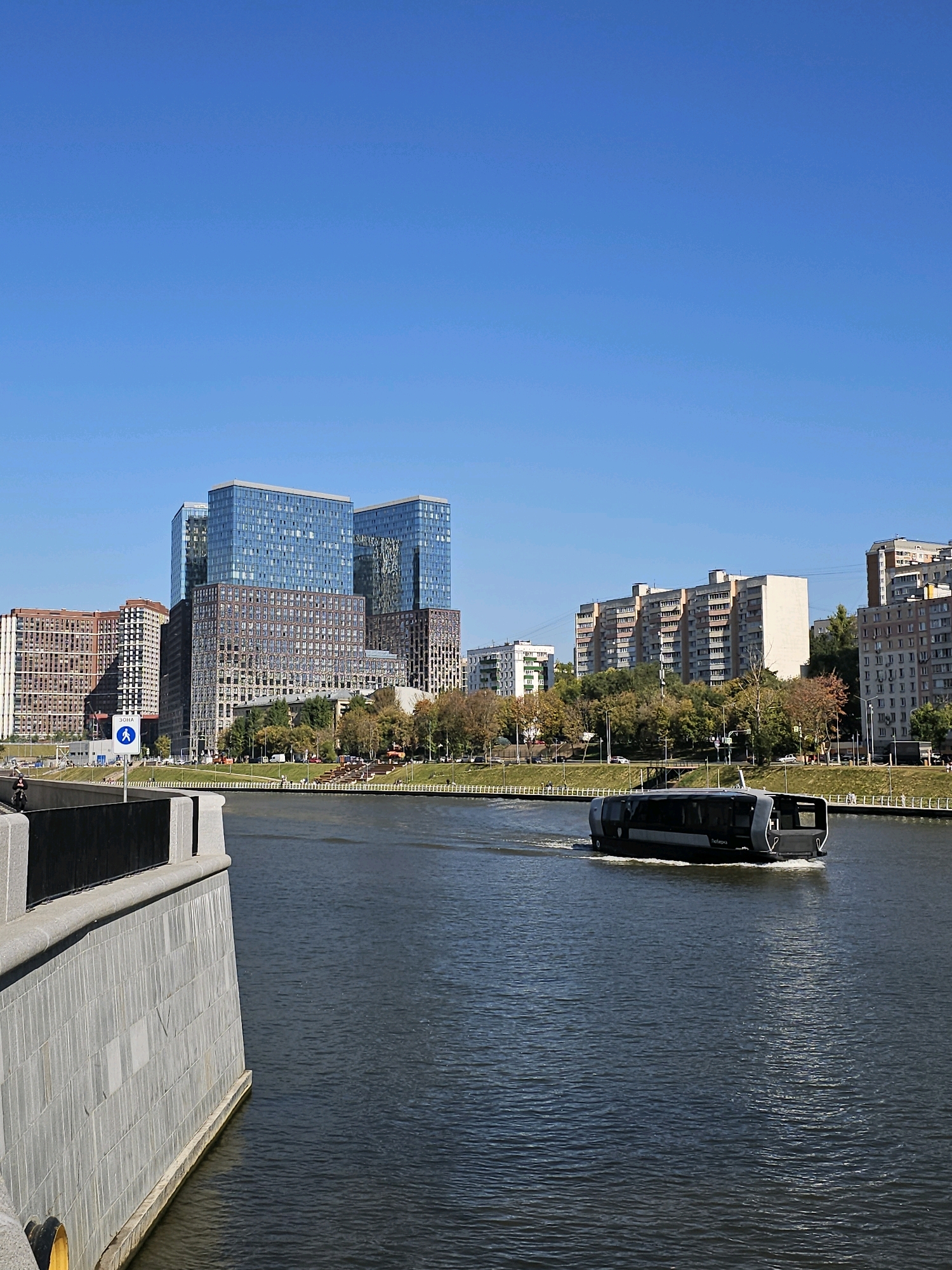 The image depicts a sunny day in Moscow, Russia, with a clear blue sky and a body of water in the foreground. On the right side of the frame, a modern, dark-colored boat with a sleek design navigates the water, leaving a white wake behind it. Across the river, a cityscape unfolds with several distinct architectural styles. To the left, a series of contemporary, glass-fronted buildings rise prominently, reflecting the sky. Adjacent to these are older, multi-story residential buildings with a more traditional facade. A tree-lined embankment separates the water from the buildings, with some greenery visible. On the left side of the image, a stone embankment with a black railing runs along the water's edge. A pedestrian crossing sign is visible near the embankment, suggesting a public area. The overall scene captures a vibrant urban environment with a blend of modern and older structures, under a bright, clear sky.