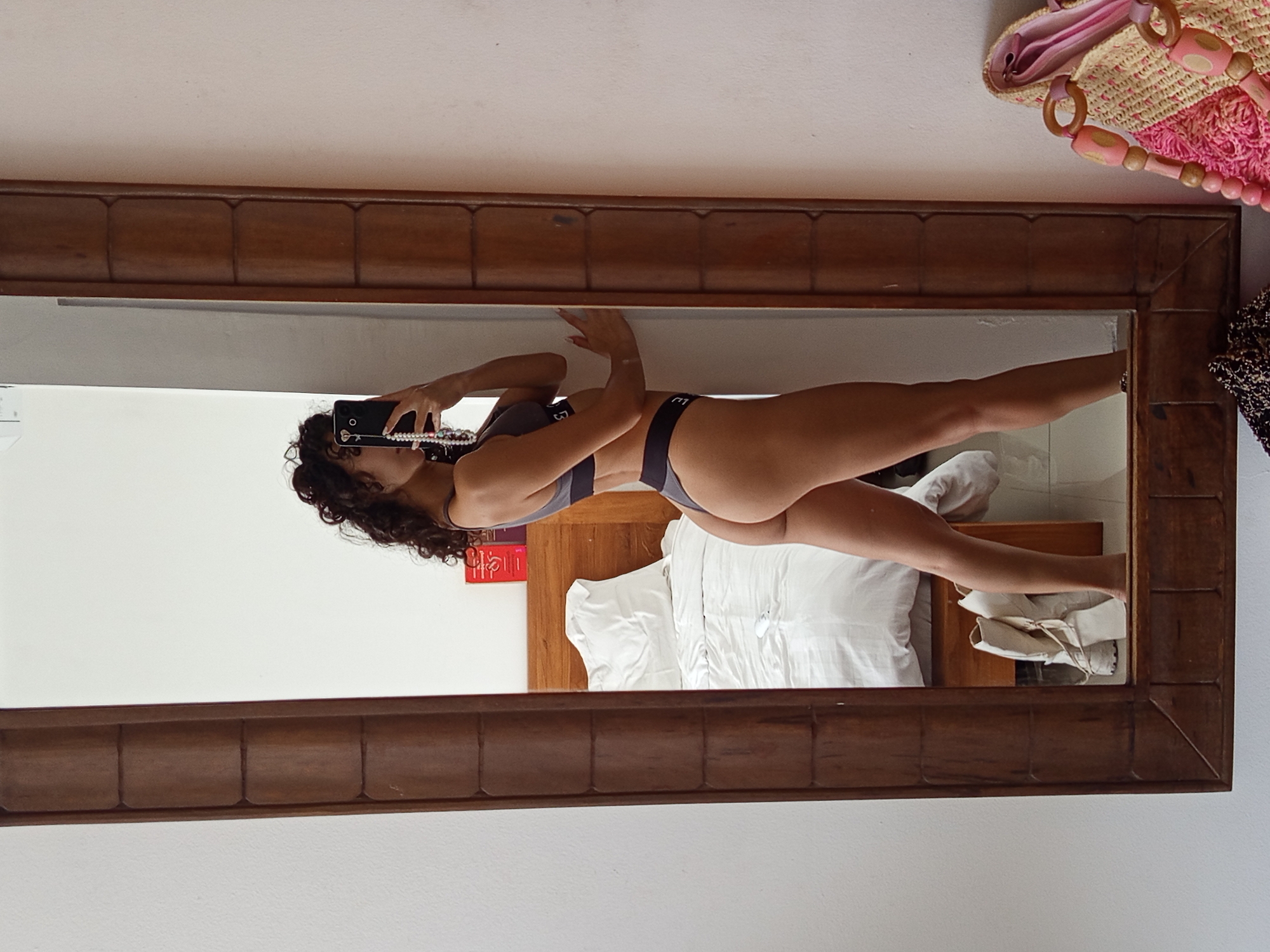 A mirrored reflection shows a woman holding a black smartphone and taking a picture of herself. She is wearing a gray bra and panties. Behind her, a bed with white linens and a headboard is visible. Next to the bed are white shoes. The photo seems to have been taken in North Kuta, Indonesia. A wooden-framed mirror captures the scene.