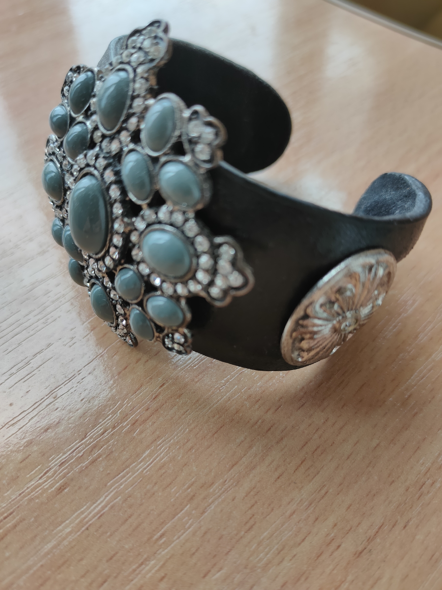 A detailed close-up shot captures a wide, black leather cuff bracelet adorned with intricate silver-toned embellishments. The central embellishment features a cluster of oval, grey-blue stones, each surrounded by smaller clear rhinestones. The stones are arranged in a symmetrical, floral-like pattern. A similar, circular silver-toned charm with a floral motif is attached to the side of the bracelet. The bracelet rests on a light brown, wooden surface with a visible grain and slight sheen, suggesting it might be a table. The lighting creates soft shadows, indicating an indoor setting. This image is not from Oktyabrsky, Russia.