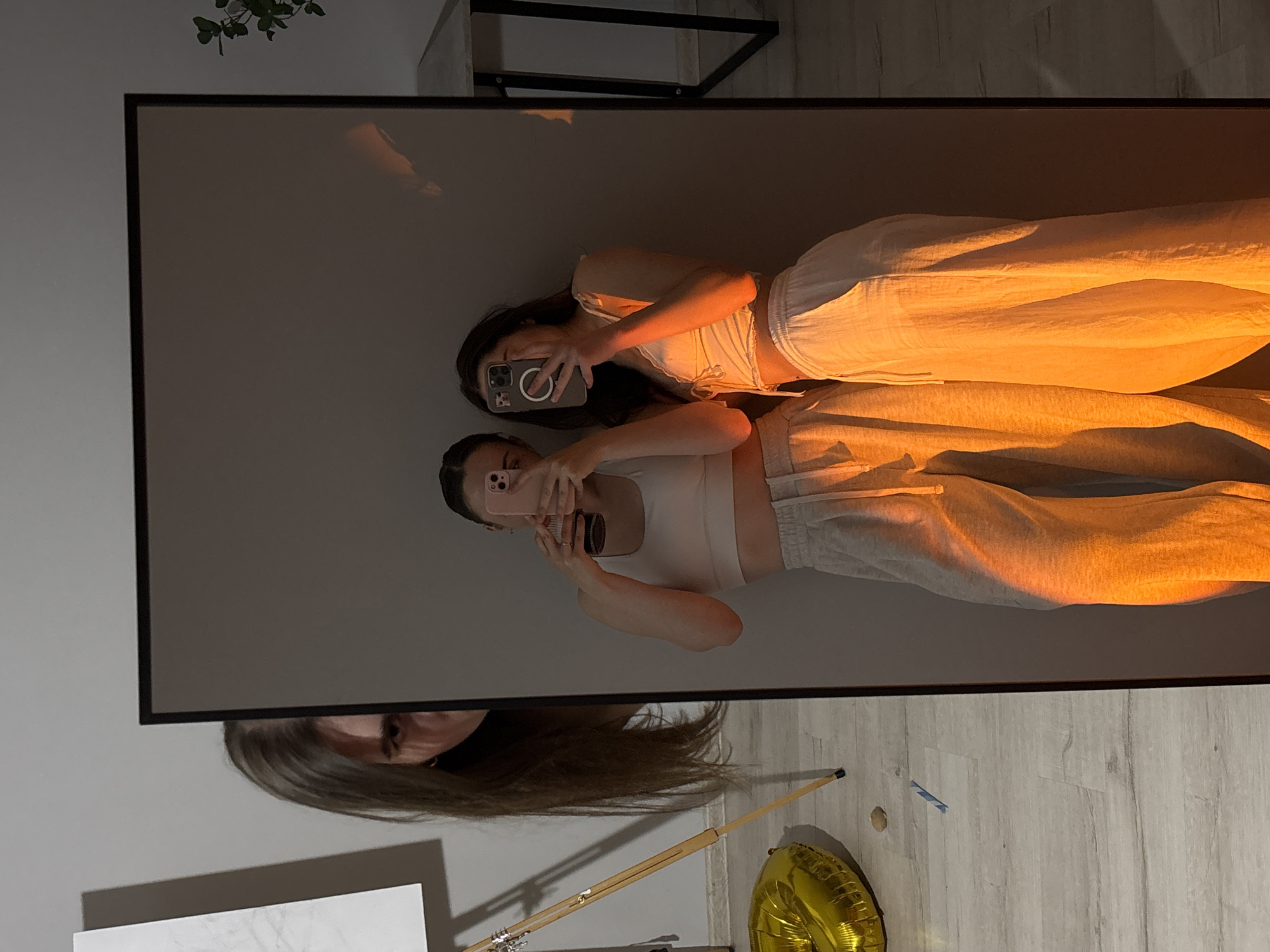 An image captures two young women taking a mirror selfie in an indoor setting. The woman on the left, partially visible, holds a pink phone, while the woman on the right, holding a dark phone, is closer to the mirror. They are both wearing casual loungewear. The lighting in the reflection is warm and orange, casting dramatic shadows. In the foreground of the actual scene, a golden balloon shaped like a number is on the floor. A tripod easel is also visible. The background shows a grey wall and a glimpse of a piece of furniture with a plant on it. The overall impression is a candid moment shared between friends. The location is specified as Ternopil, Ukraine.