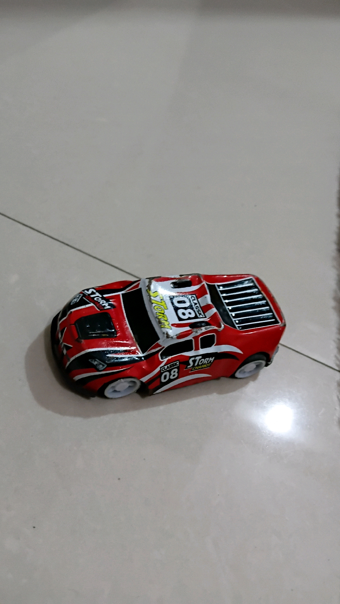 A red and white toy race car is positioned on a tiled floor. The car has black accents and features the number "08" and the word "STORM" printed on its sides and roof. The car is facing left and is slightly angled towards the viewer. The background consists of light-colored square tiles. The image was taken in the city of Klorogan, Indonesia.