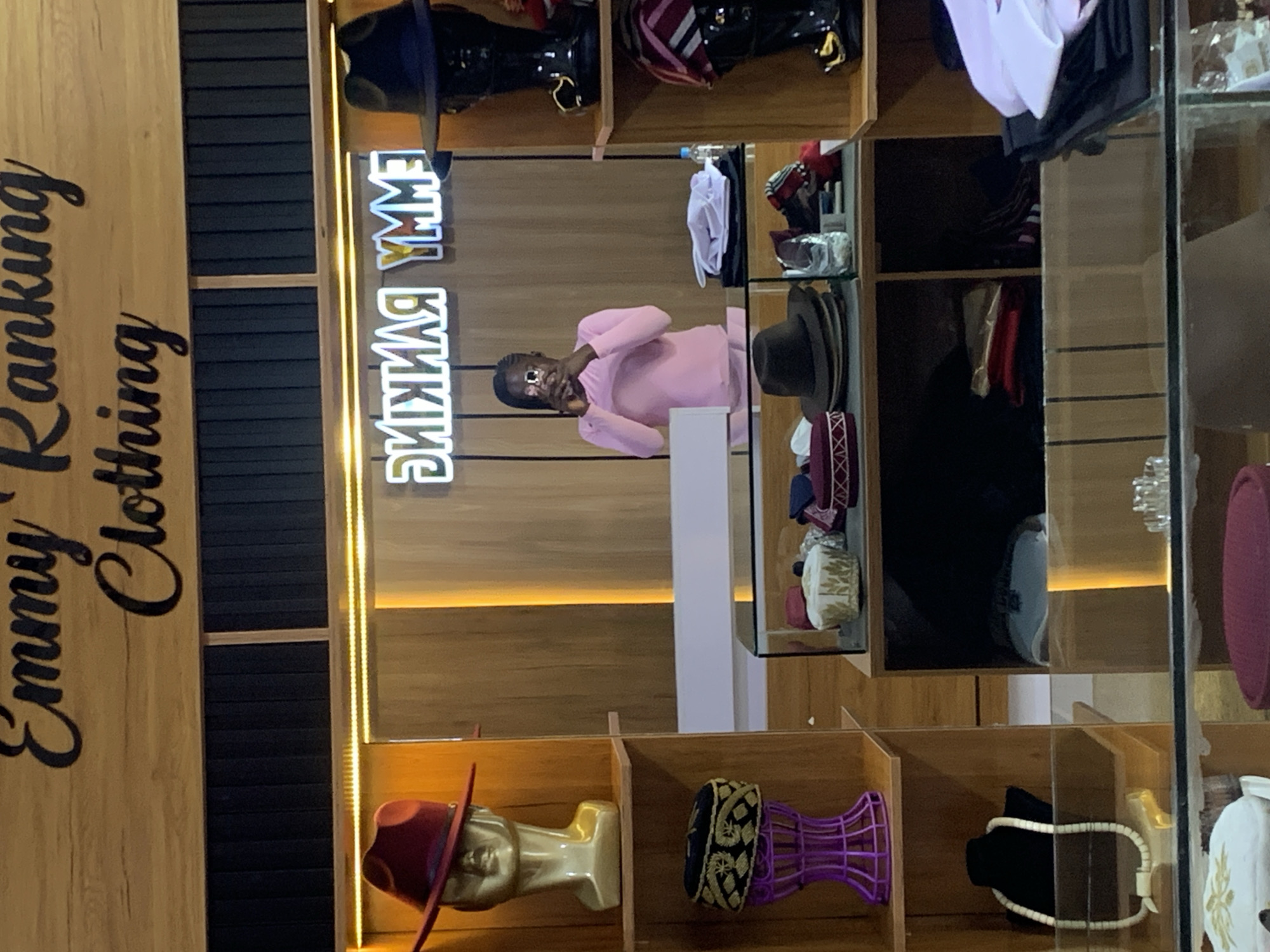 The image shows the interior of a clothing store. A neon sign with the text "EMMY RANKING" is visible on the wall. The store appears to be well-stocked with hats and other accessories. In the foreground, there is a display case filled with various hats. On the shelves to the left, there are mannequins wearing hats and a necklace. A woman is standing behind a counter, taking a picture with her phone. The store's branding, "Emmy Ranking Clothing," is displayed prominently above the entrance. This scene is set in Gura Top, Nigeria.