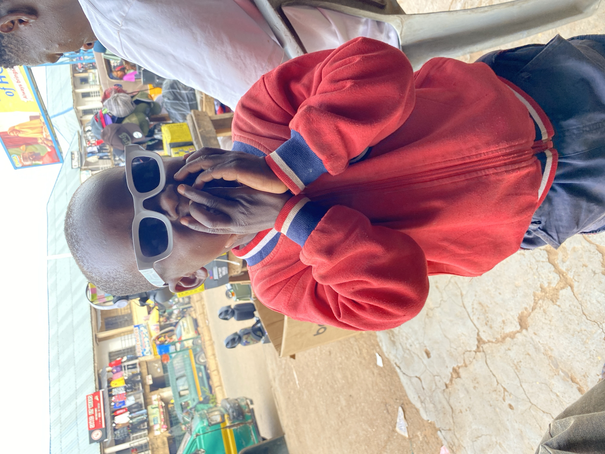 A photograph taken from a low angle depicts a child wearing white-rimmed sunglasses and a red zip-up jacket with blue, white, and red striped cuffs. The child has dark skin and a shaved head, and their hands are partially covering their face. The background reveals an outdoor market scene in Bukuru, Nigeria, with various commercial establishments and vendors. People are visible amidst a cluttered street scene with shops, vehicles, and goods on display. A partial view of an adult is seen in the upper part of the frame. The ground appears to be cracked concrete.