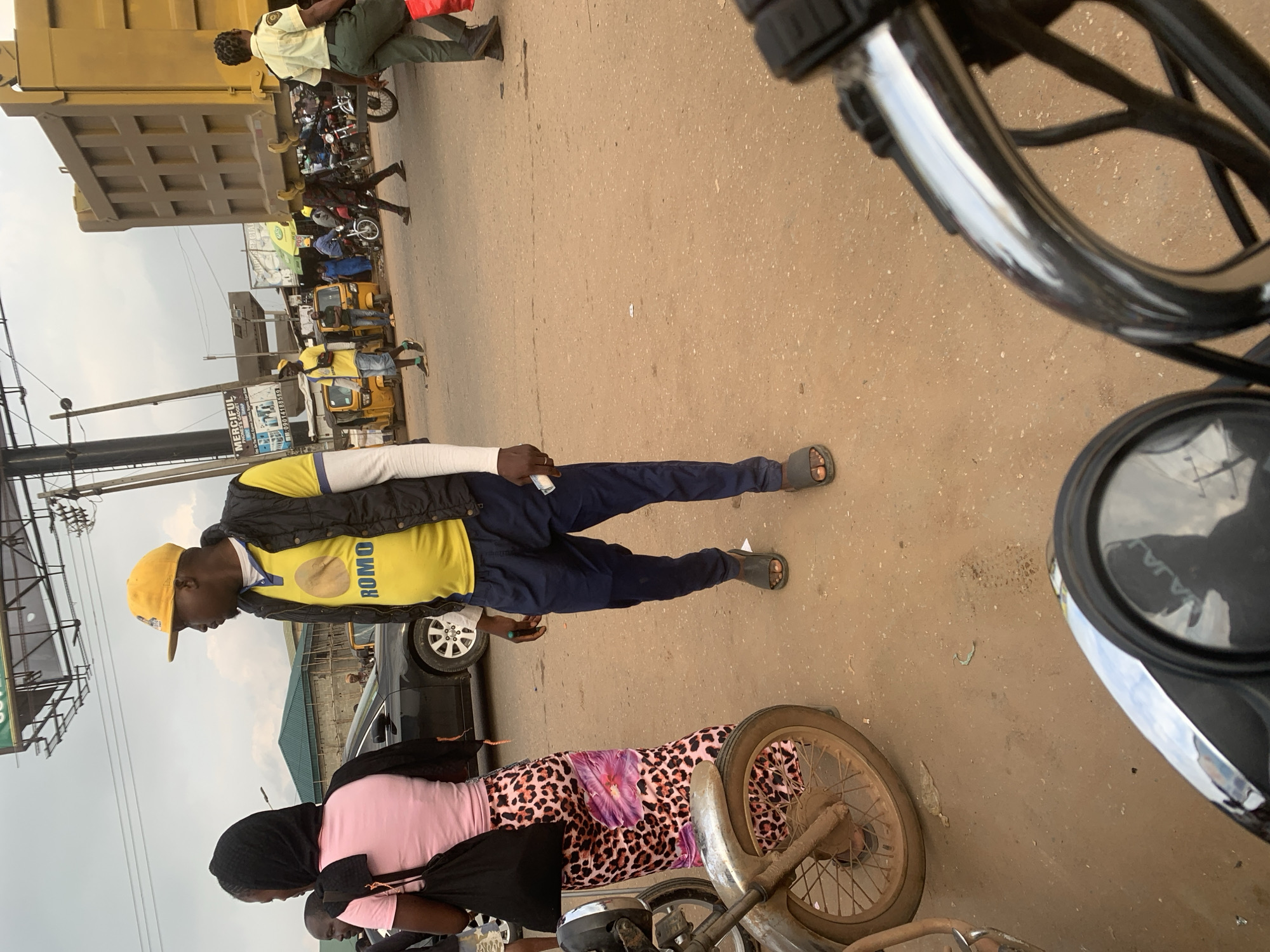 In Shagamu, Nigeria, on a dusty, unpaved street, a man wearing a yellow cap, yellow polo shirt with "ROMO" printed on it, and a dark vest walks towards the right. He is wearing dark blue pants and dark sandals. To his left, a woman wearing a pink shirt and a black headscarf also faces right, carrying a black bag. A motorcycle is parked to the left of the frame. In the background, a large yellow truck is parked, and other people and vehicles, including tricycles and motorcycles, are visible on the street. The sky is overcast.