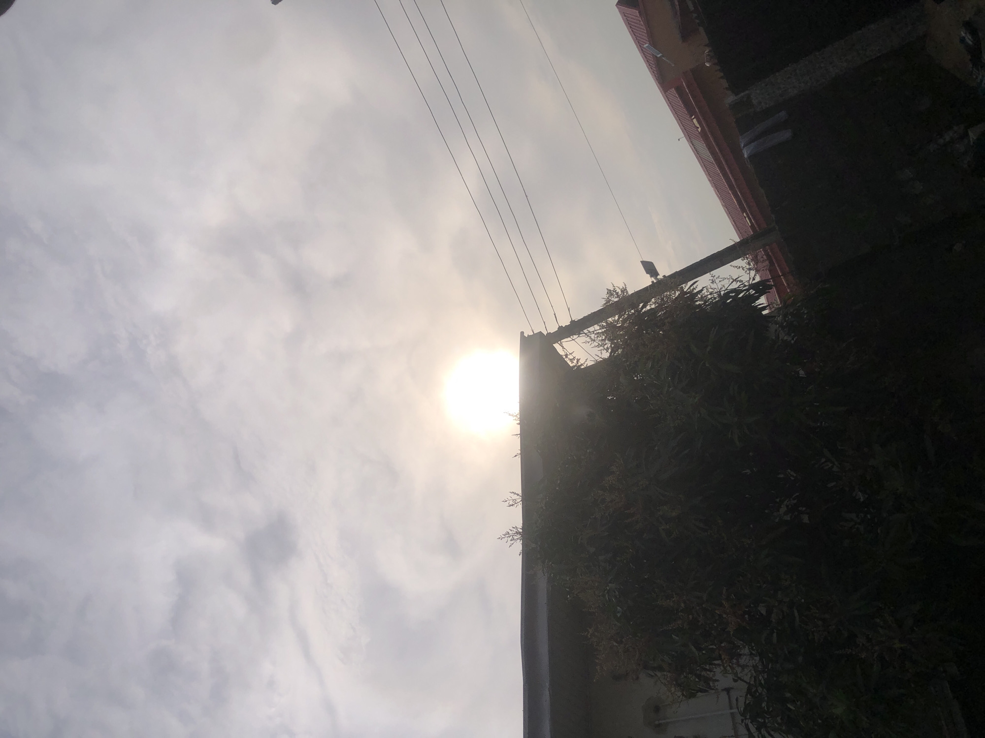The image captures a view of a sky with clouds and the sun partially obscured. In the foreground, the dark silhouette of a tree with some visible branches and foliage is present, along with the edge of a building with a dark roof. Another building with a red roof is visible in the background, along with a wall and some hanging clothes. Power lines are stretched across the sky. The scene appears to be an outdoor setting, possibly in a residential area of Ado, Nigeria, given the architectural style of the buildings and the presence of overhead power lines. The lighting suggests it could be late afternoon or early morning. No people are explicitly visible.