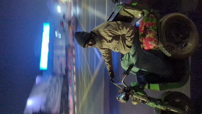 A man with a dark beanie and tan jacket sits on a green motorized rickshaw, likely a tuk-tuk. He wears gloves and has a small beard. The vehicle has a seat behind him and a covered wheel. A building with a bright blue neon sign is in the background, blurred with motion. The scene appears to be at night, possibly in a city, with wet pavement reflecting light.