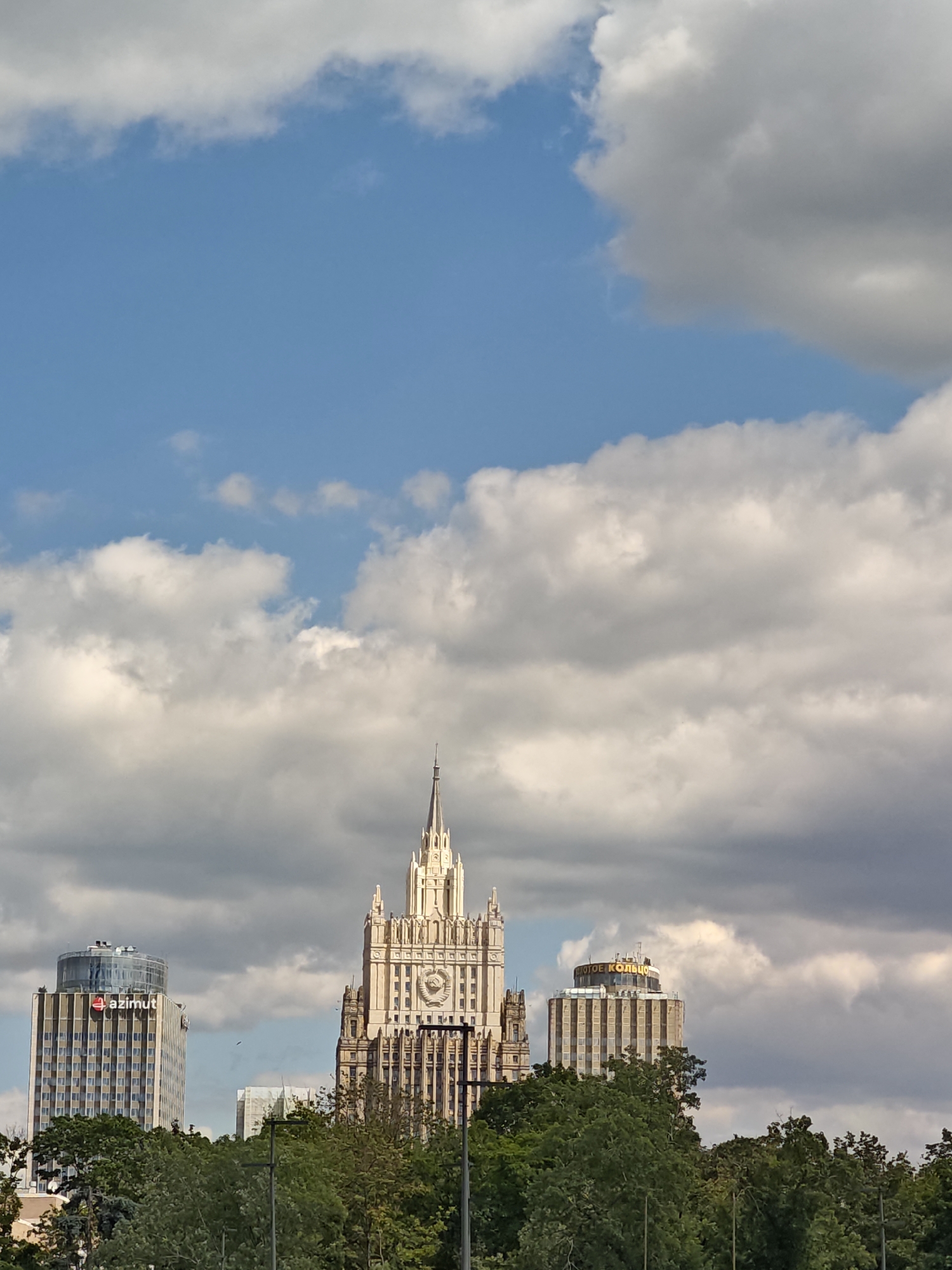 The image shows a cityscape with several tall buildings under a partly cloudy blue sky. In the center is a prominent, ornate Stalinist-era skyscraper topped with a spire, featuring a large clock face and a Soviet star emblem. To the left of this central building is a modern high-rise with the "AZIMUT" logo on its upper section. To the right, another building with a rounded top displays text in Russian, likely a hotel name. A line of lush green trees forms the foreground, obscuring the lower parts of the buildings and street level. The overall scene captures a mix of architectural styles in an urban environment. This view is likely of the Ministry of Foreign Affairs building and surrounding structures on Smolenskaya Square in Moscow, Russia.