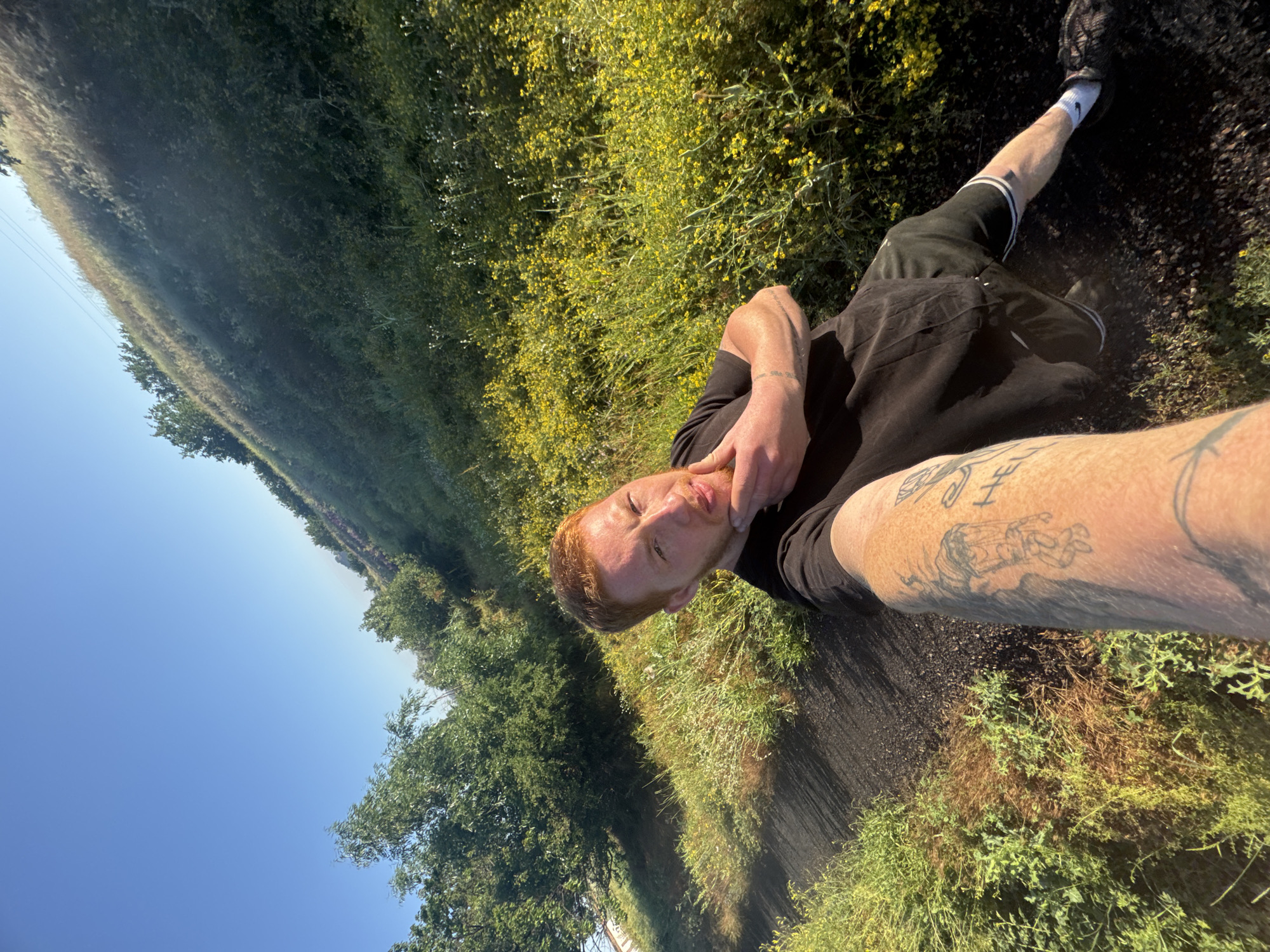 This media file is a selfie taken outdoors on a sunny day. The subject is a person with red hair and fair skin, wearing a black t-shirt and black shorts with white stripes. They have tattoos on their arm. The person is looking at the camera, with their hand resting on their chin, appearing to be relaxed or pensive.
The background consists of lush green vegetation, including trees and flowering plants with small yellow blossoms, suggesting a natural or rural setting. A clear blue sky is visible in the upper left portion of the image. The ground appears to be dark and gritty, possibly a dirt path or road. The overall impression is that of a person enjoying a warm day outdoors, perhaps on a walk or hike. The lighting indicates it is daytime, likely late morning or early afternoon due to the bright sunlight and shadows. No text is visible in the image.