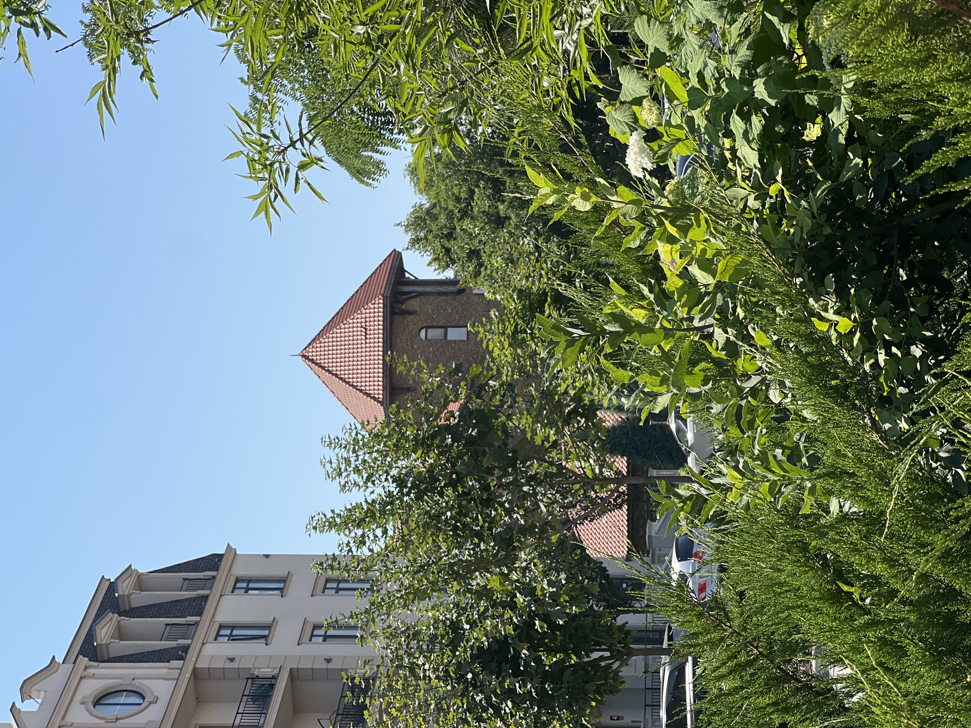 The image depicts a sunny outdoor scene in Fontanka, Ukraine, with a clear blue sky. In the foreground, dense green foliage, including coniferous trees and leafy bushes, partially obscures the view. Behind the greenery, a modern cream-colored apartment building with a mansard roof and dark windows stands to the left. To the right of the apartment building, a reddish-tiled tower with small windows and wooden accents rises amidst more trees. Several cars are parked on a street visible between the apartment building and the tower. The overall impression is one of an urban environment interspersed with lush vegetation.