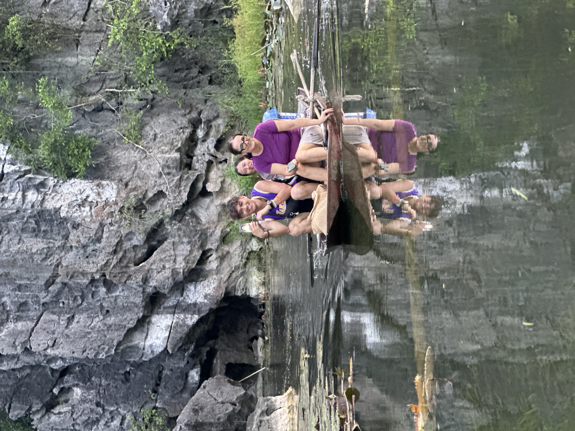 Three individuals are in a small boat navigating through a body of water surrounded by limestone karsts. The person in the front left, a young male wearing a purple and yellow tank top, is holding up a smartphone, seemingly taking a photo. He is sitting next to a straw hat. To his right sits a young female with dark hair, smiling. The third person, a woman wearing a purple shirt and beige shorts, is at the back of the boat, holding a phone in her hand. She is looking forward. The water's surface reflects the boat and its occupants. The background features rugged, grey rock formations with sparse vegetation and some water lilies. The scene appears to be a natural landscape, likely a tourist attraction in Nam Hoa Lư Ward, Vietnam, known for its karst topography and waterways.