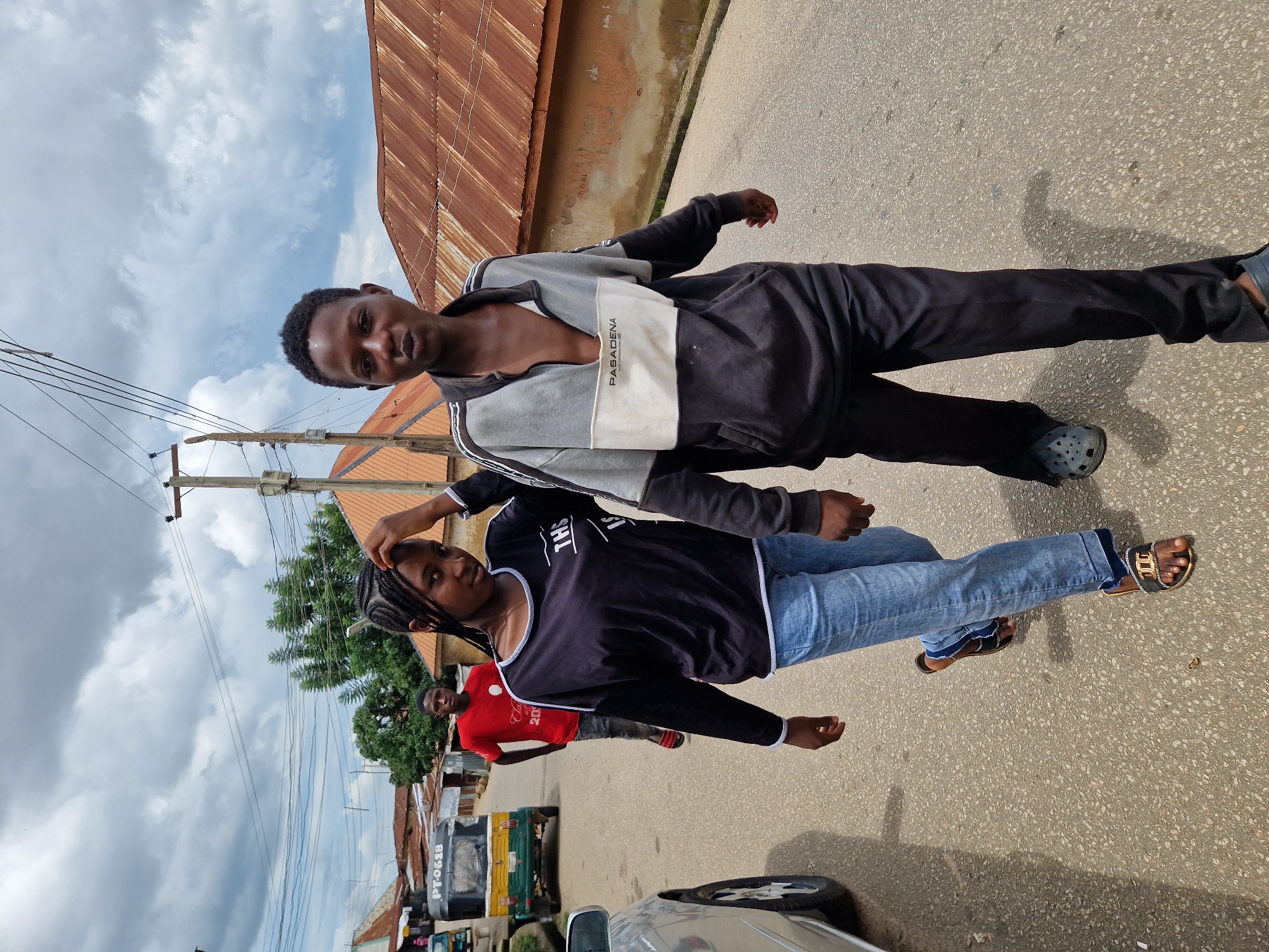 In an outdoor, street-level view under a cloudy sky, two young individuals, a female and a male, walk towards the camera on a paved road in a residential area. The female, on the left, wears a black long-sleeved shirt with white trim and blue jeans, accessorized with elaborate sandals. Her hair is styled in braids. The male, on the right, is dressed in a grey and dark-colored zip-up jacket with white trim and black pants, paired with grey clogs. He is making a puckering gesture with his lips. In the background, a man in a red t-shirt stands further down the street. Several buildings with corrugated metal roofs line the road, and a three-wheeled vehicle is visible on the left. The environment appears to be a modest urban or semi-urban setting.