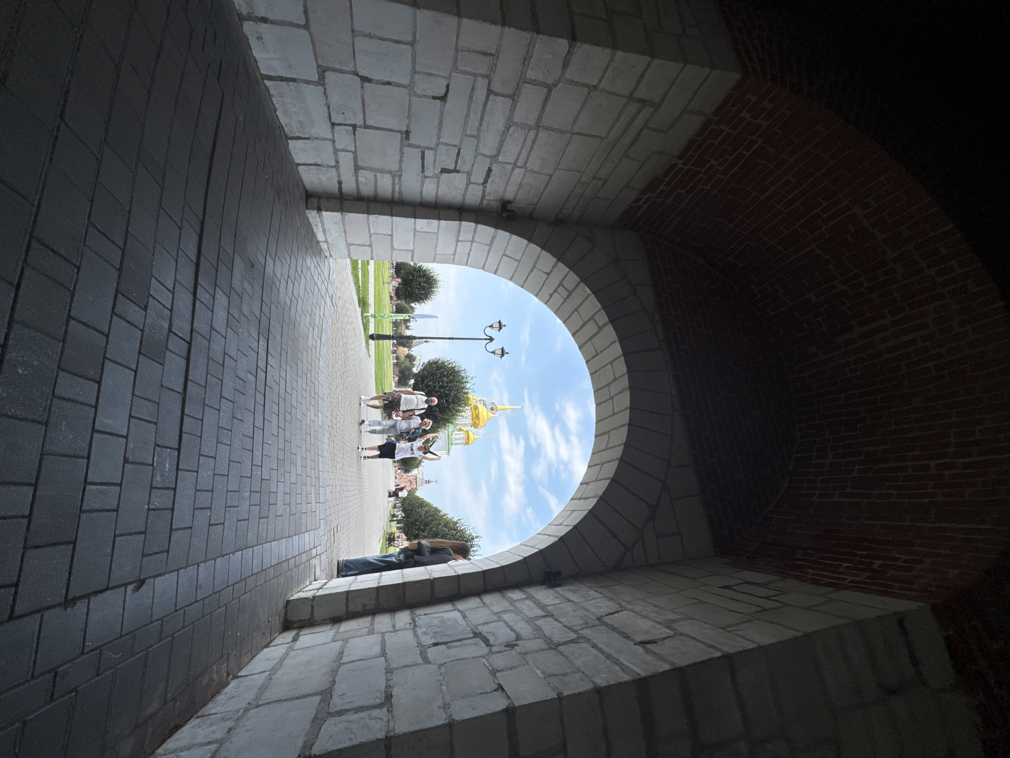 The image captures a view from within a large stone archway, looking out onto a sunny outdoor plaza. The archway is constructed with large, light-colored stone blocks on the sides and a dark brick-lined ceiling. The ground within the archway is paved with dark grey rectangular stones.
Through the arch, a paved walkway leads into a green park-like area. Several people are visible on the walkway and in the park. In the mid-ground, a group of four people are walking towards the viewer. Two are adult males, one is an adult female, and one is a child. The child in the center of the group has their arms raised. Further in the distance, a tall, black lamppost with two lights stands on the left side of the walkway. Beyond the lamppost, several trees with rounded canopies are planted in rows. In the far background, prominent golden domed buildings are visible against a bright blue sky with scattered white clouds.
The scene is likely situated in the Tula Kremlin, a historic fortress in Tula, Russia, known for its red brick walls and golden-domed churches. The architectural style of the archway and the presence of the golden domes strongly suggest this location. The overall atmosphere is bright and pleasant, indicating a warm day.