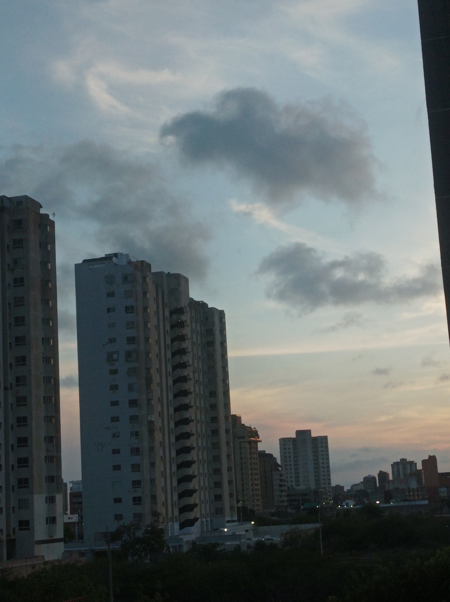 The image shows a cityscape at dusk. Several tall apartment buildings are visible, lining the left side of the frame and extending into the distance. The buildings appear to be constructed of concrete and have numerous windows. In the background, there are additional buildings of varying heights, also appearing to be residential or commercial structures. The sky is filled with clouds, with a gradient of colors from blue to orange near the horizon, indicating the setting sun. There is greenery visible at the bottom of the frame, suggesting the presence of trees or vegetation. A dark, undefined object is present on the right side of the image. The location is Unknown, Unknown.