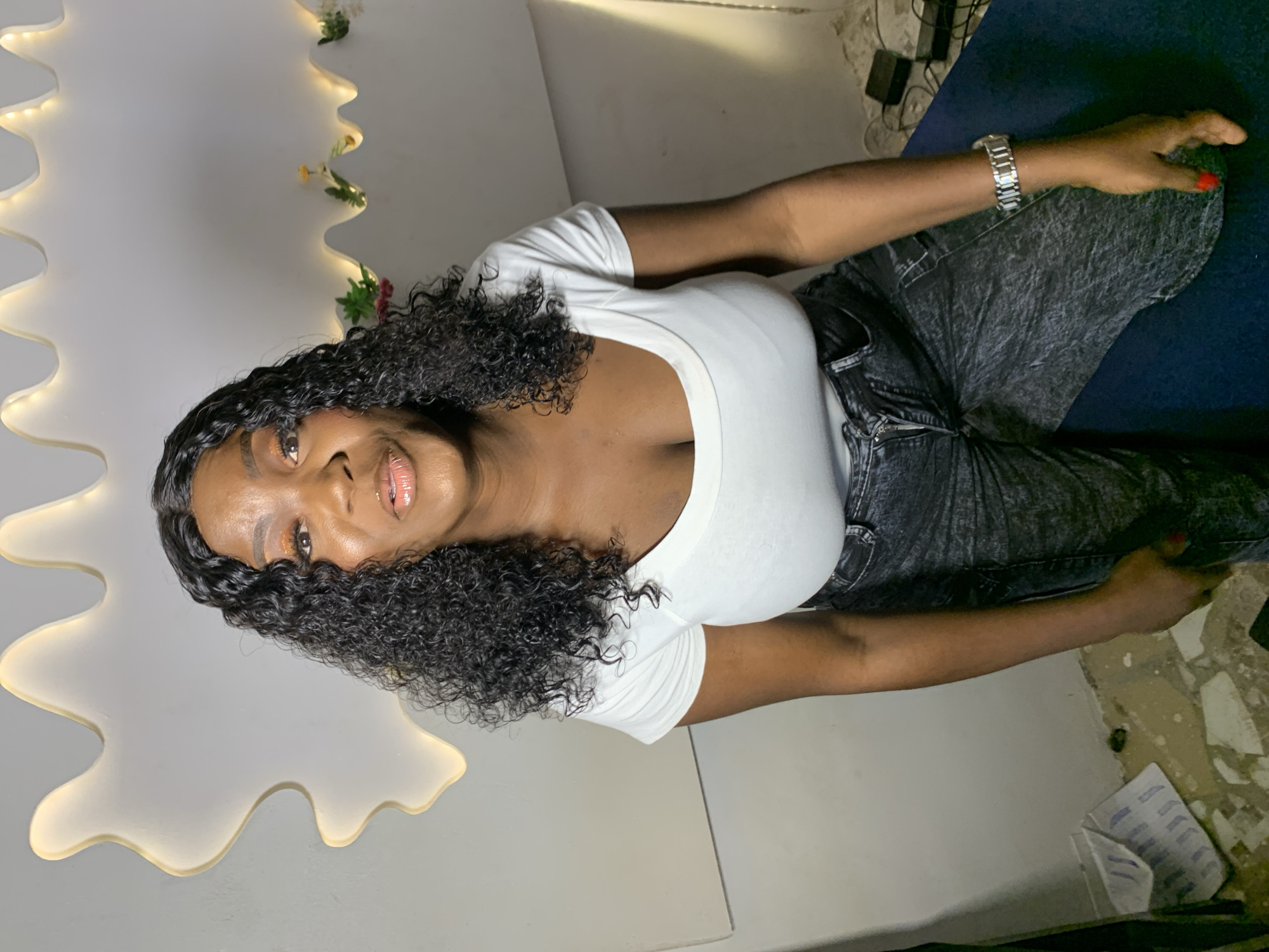 A person, likely female, is depicted in a portrait. The subject has dark, curly hair and is wearing a white t-shirt and dark, possibly denim, pants. She is adorned with makeup, including eyeshadow and lip gloss, and a silver watch on her left wrist. Her fingernails are painted red. She is positioned in a studio-like setting with a white, irregular-shaped backdrop featuring embedded lights and some small artificial floral decorations. The floor appears to be tiled and somewhat worn. The lighting in the scene is artificial, casting soft illumination. The overall impression is that of a casual, posed photograph, possibly taken indoors. The background elements like the lights and the smooth, abstract shape of the backdrop suggest a deliberate setup for photography or videography. There is no visible text in the image.