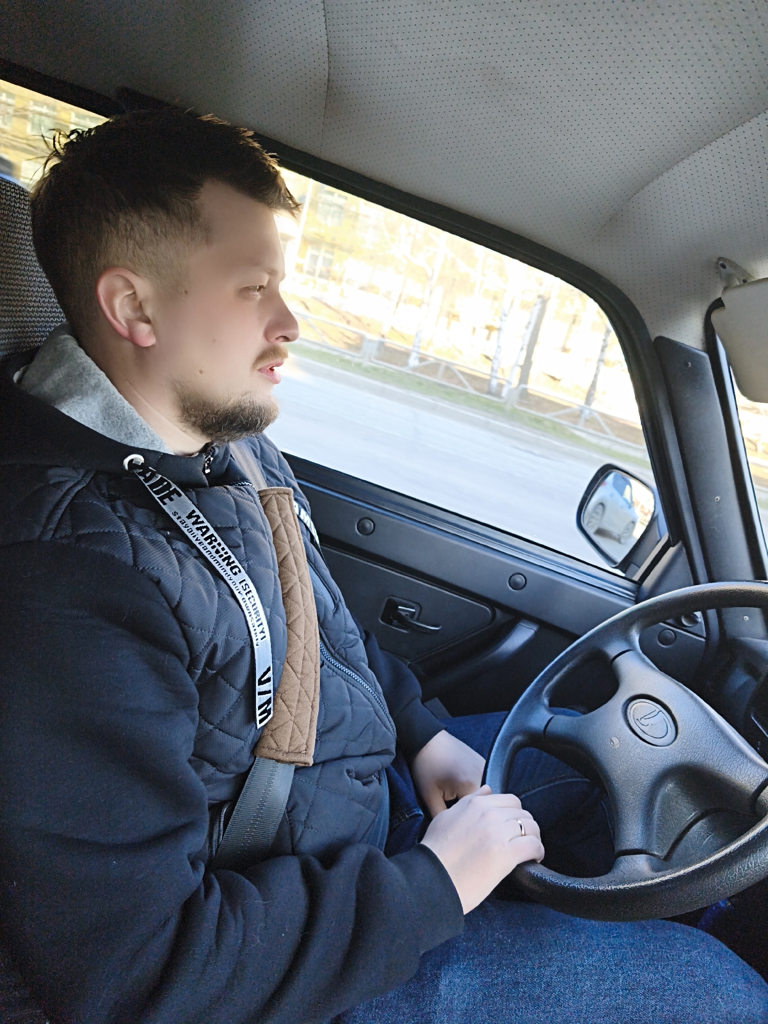 A man is driving a car. He is wearing a black jacket with a gray hoodie underneath and blue jeans. He has a short haircut and a beard. The car's steering wheel is black and has a logo in the center. The car is moving along a city street. The location is Unknown, Unknown.