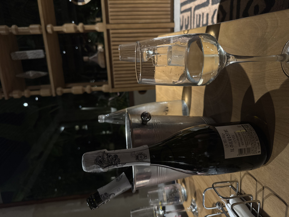 A tilted shot showcases a bottle of "Il Silvestri Cuvee Extra Dry" sparkling wine in a silver ice bucket. The bucket contains empty bottles. A partially filled wine glass rests nearby on a wooden surface. The background includes a wooden shelf and glimpses of greenery outside, creating an ambiance of indoor/outdoor dining or a bar setting.