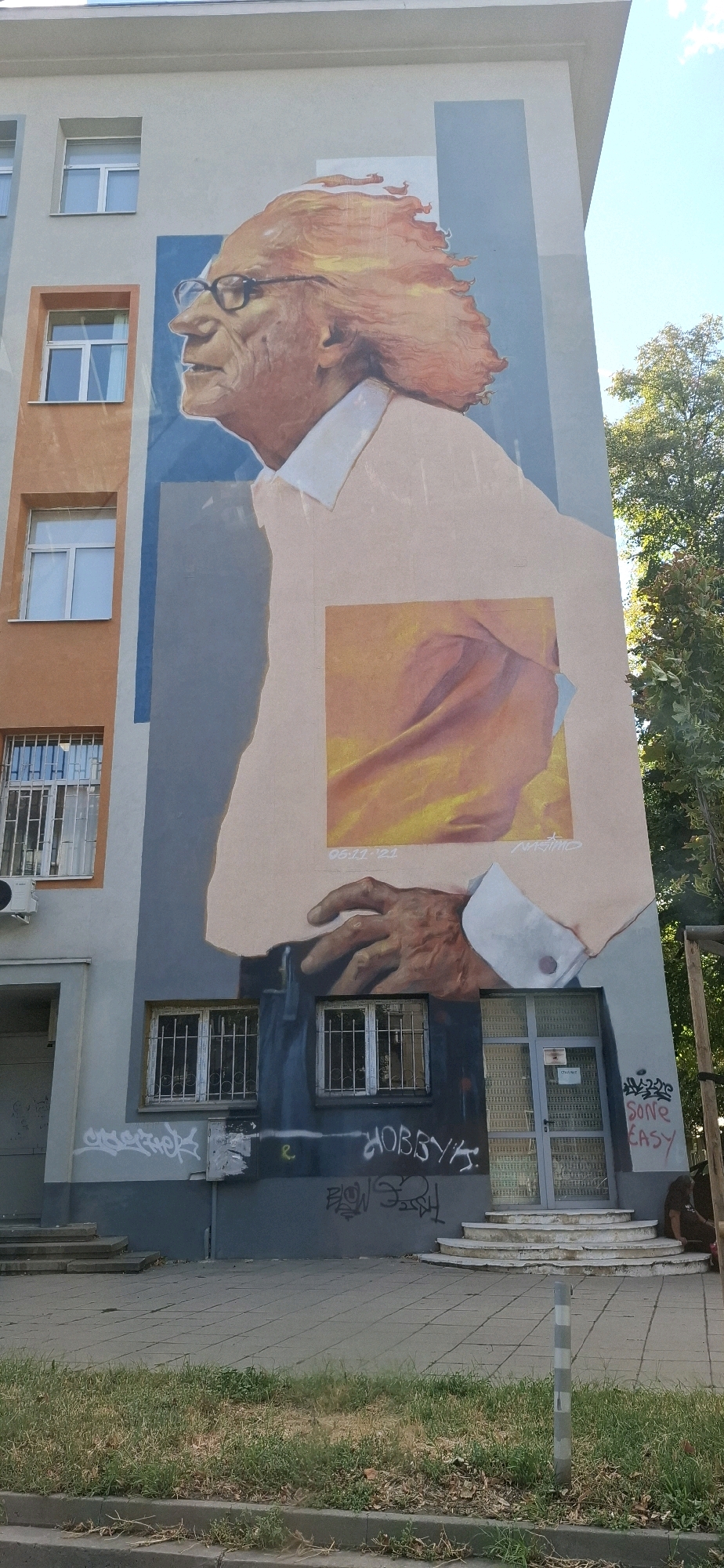 This is a street-level, eye-level, full-shot photograph of a building with a large mural on its side, located in Sofia, Bulgaria. The mural depicts a close-up portrait of an elderly man with prominent glasses and wild, orange hair. He is wearing a light-colored shirt and a dark suit jacket. The mural extends from the top of the building down to the first floor. Below the mural, on the first floor of the building, there are several windows and an entrance with a glass door. The building's exterior is painted in muted tones of gray, light blue, and orange. The foreground shows a sidewalk paved with square tiles, a strip of dry grass, and a curb. There is graffiti on the lower part of the building's facade. The sky above is a clear blue.
