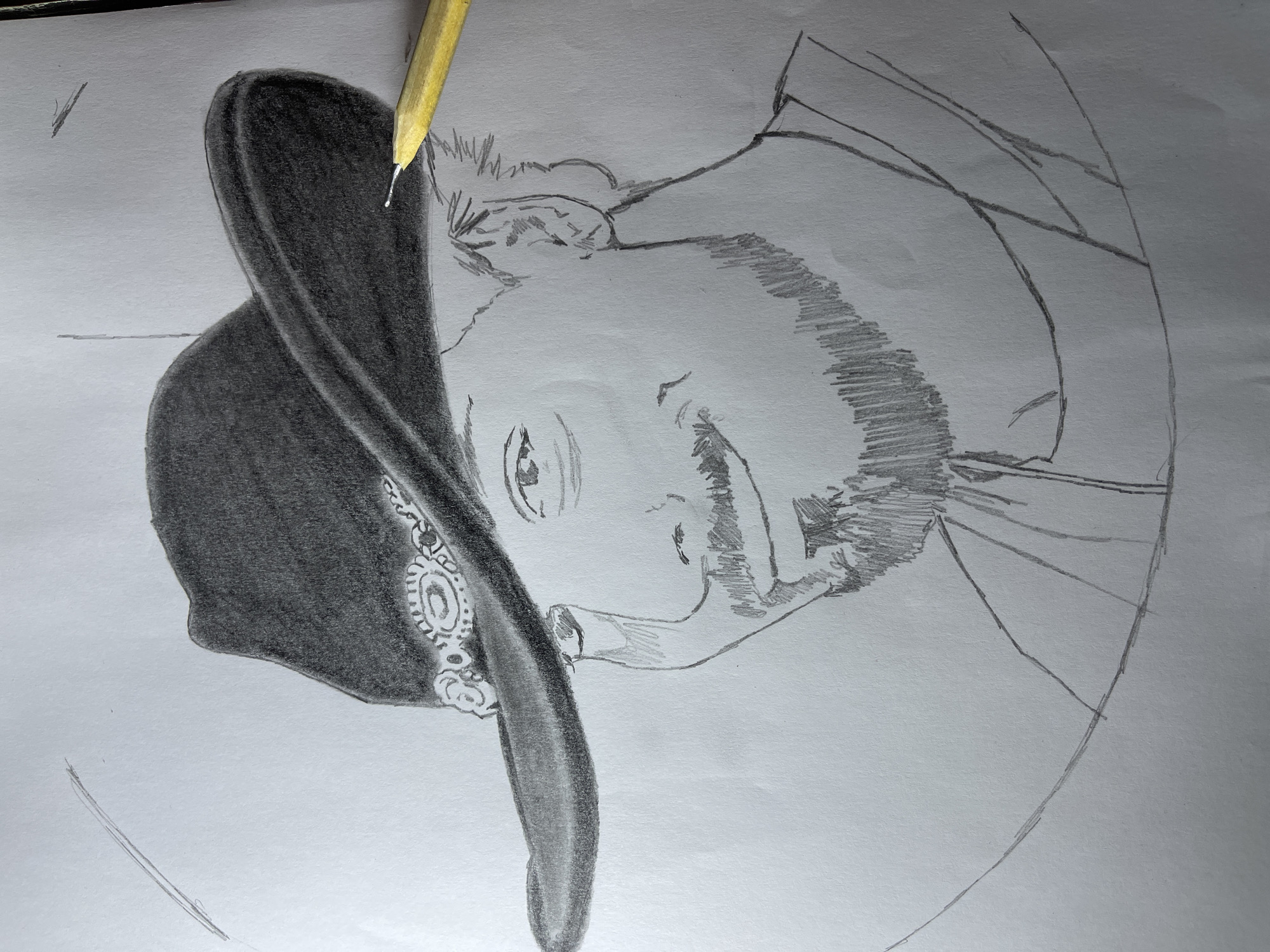 A black and white pencil drawing on white paper depicts a man with a hat and beard. A yellow pencil rests atop the hat. The background appears to be plain. The drawing is oriented upside down in the frame. No location in Jos, Nigeria, is evident.
