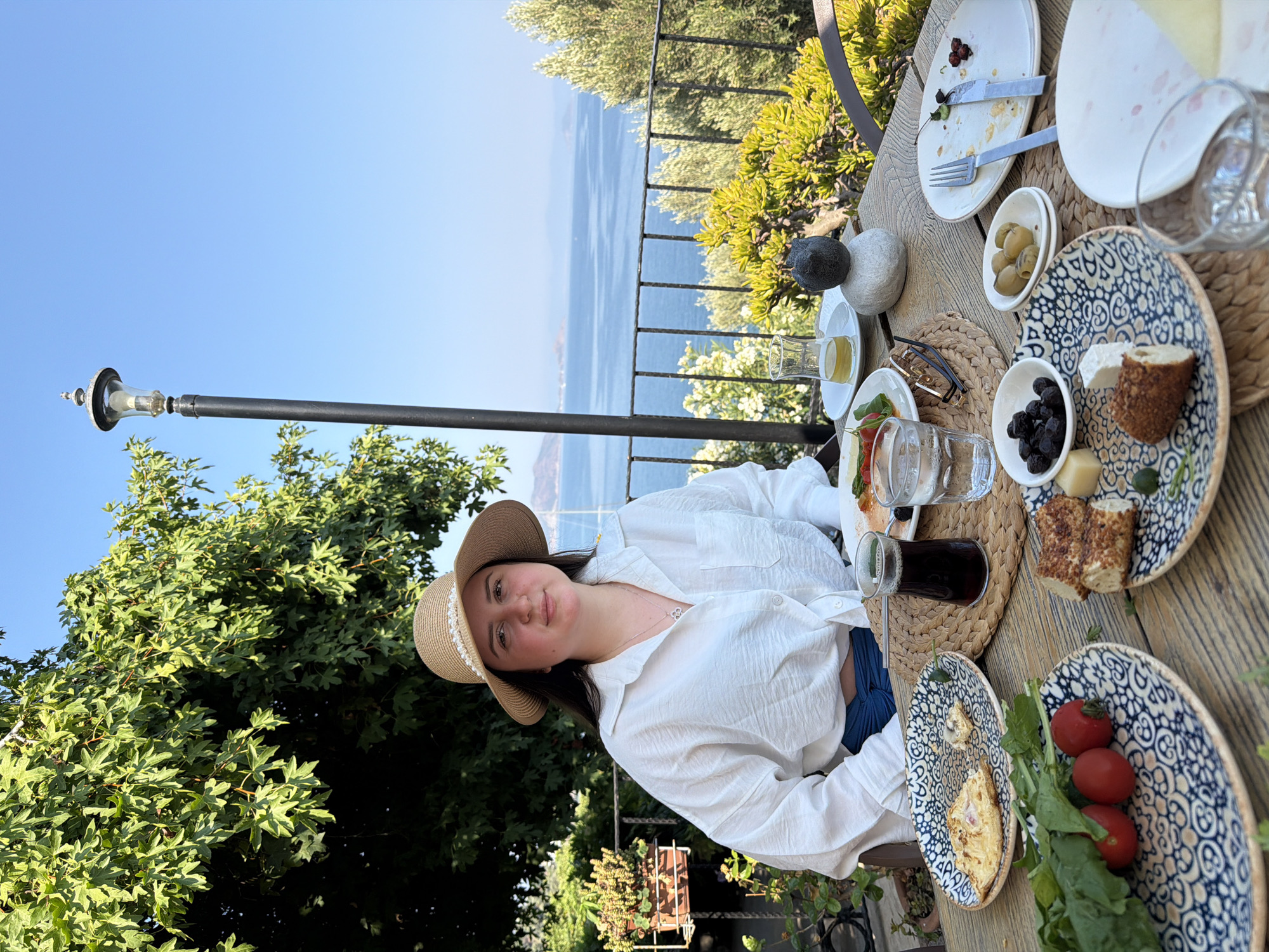 A young woman wearing a straw hat and a white linen shirt is seated at a wooden table outdoors. The table is set with plates of food, including pastries, olives, tomatoes, and greens. A glass of dark liquid and a glass of water are also on the table. The woman is looking at the camera with a slight smile. In the background, a body of water with distant hills can be seen. The scene appears to be in a restaurant or cafe with an outdoor seating area overlooking the sea, likely in Fethiye, Turkey, given the Mediterranean landscape. The woman's attire and the setting suggest a relaxed, daytime meal.