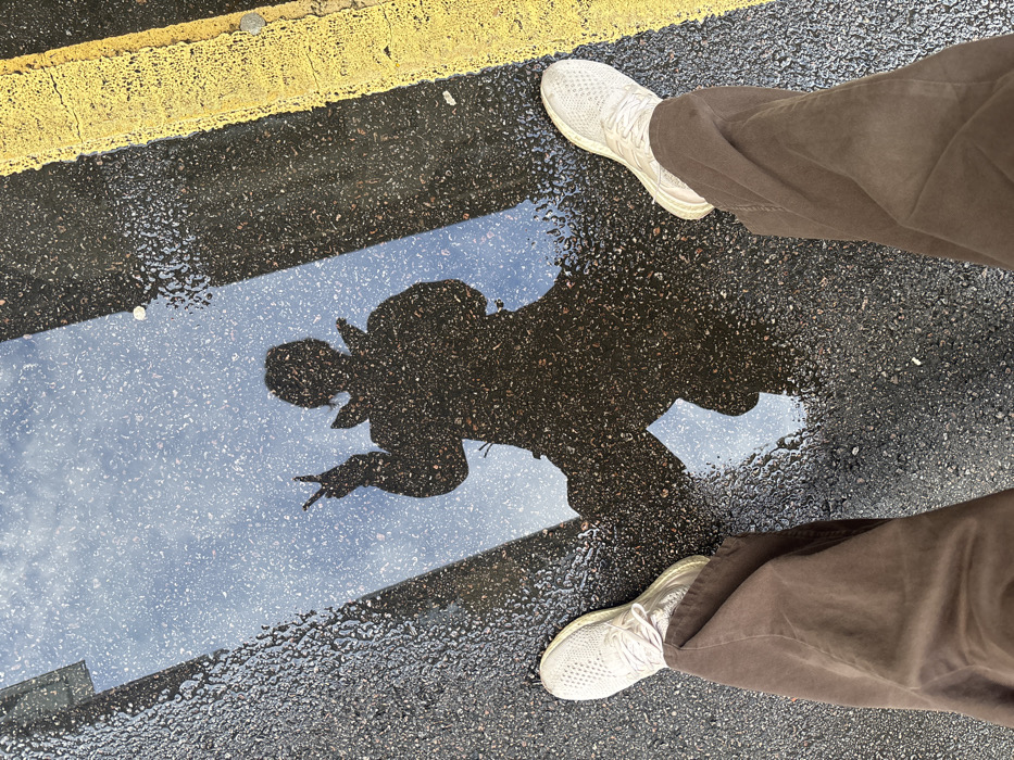 The image captures a first-person, downward-facing perspective on a wet asphalt surface in the City of London, United Kingdom. The bottom of the frame shows a person's legs, clad in brown, wide-leg trousers and light-colored, textured athletic shoes. Adjacent to the footwear is a large, rectangular puddle reflecting a light blue sky with indistinct cloud formations. Prominently reflected within the puddle is the dark silhouette of a standing individual, presumed to be the photographer, wearing a coat or jacket. The silhouette's left hand is raised, displaying a "peace sign" with two extended fingers. To the right of the puddle, two parallel, slightly faded yellow lines are visible on the asphalt. The surrounding ground appears damp, indicating recent precipitation.