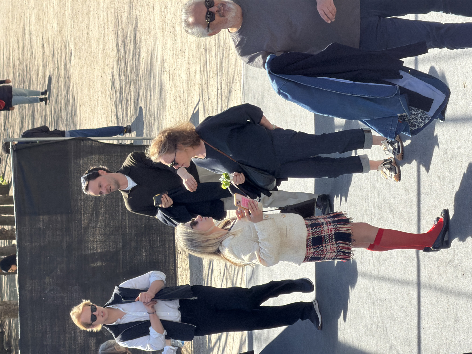 The image depicts a group of people standing outdoors on a paved surface. The scene appears to be taking place in a public area, possibly in the city of Paris, France.
Several individuals are visible, including a woman in a white jacket and plaid skirt, holding a pink smartphone. Another woman is holding a small bouquet of white flowers. Some individuals are looking at their phones.
The background includes a dark, possibly temporary structure and other people. The overall environment suggests a casual, urban setting.