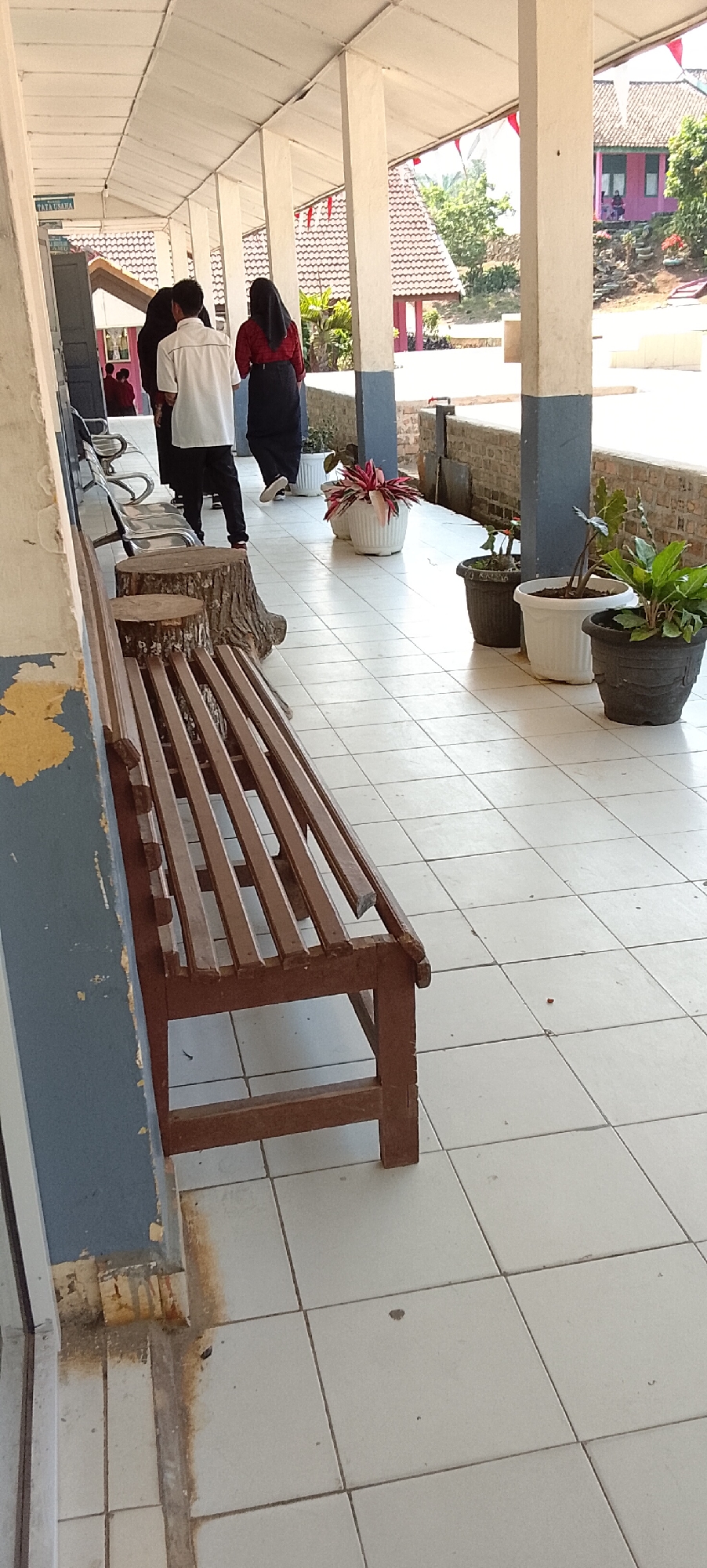 The image depicts a hallway with tiled flooring, featuring a wooden bench in the foreground and several potted plants lining the wall. Three individuals are walking away from the camera down the hallway. The architecture suggests a public building, possibly a school or administrative office, with a red-tiled roof visible in the background. The context of Jepara, Indonesia, implies a tropical climate, which is consistent with the lush greenery seen outside. The overall scene is mundane and captures a moment of everyday life in an outdoor covered walkway.