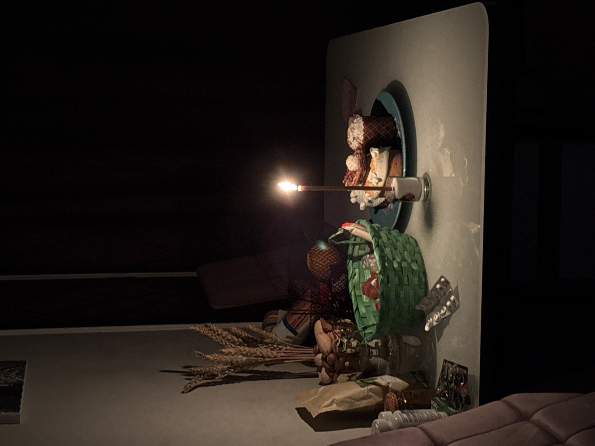 The image depicts a dimly lit indoor scene. A white, rounded-edge board is positioned against a dark background. On the board, a light-blue plate holds stacked food items and a white cup. A lit match or toothpick is held up to the food, providing the primary light source. Below the board, a small pile of objects including wheat, nuts, and packaged goods sit on what appears to be a flat surface. A green decorative basket is also visible. The background is dark, with a faint rectangular shape visible. The scene is indoors. The location is Unknown, Unknown.