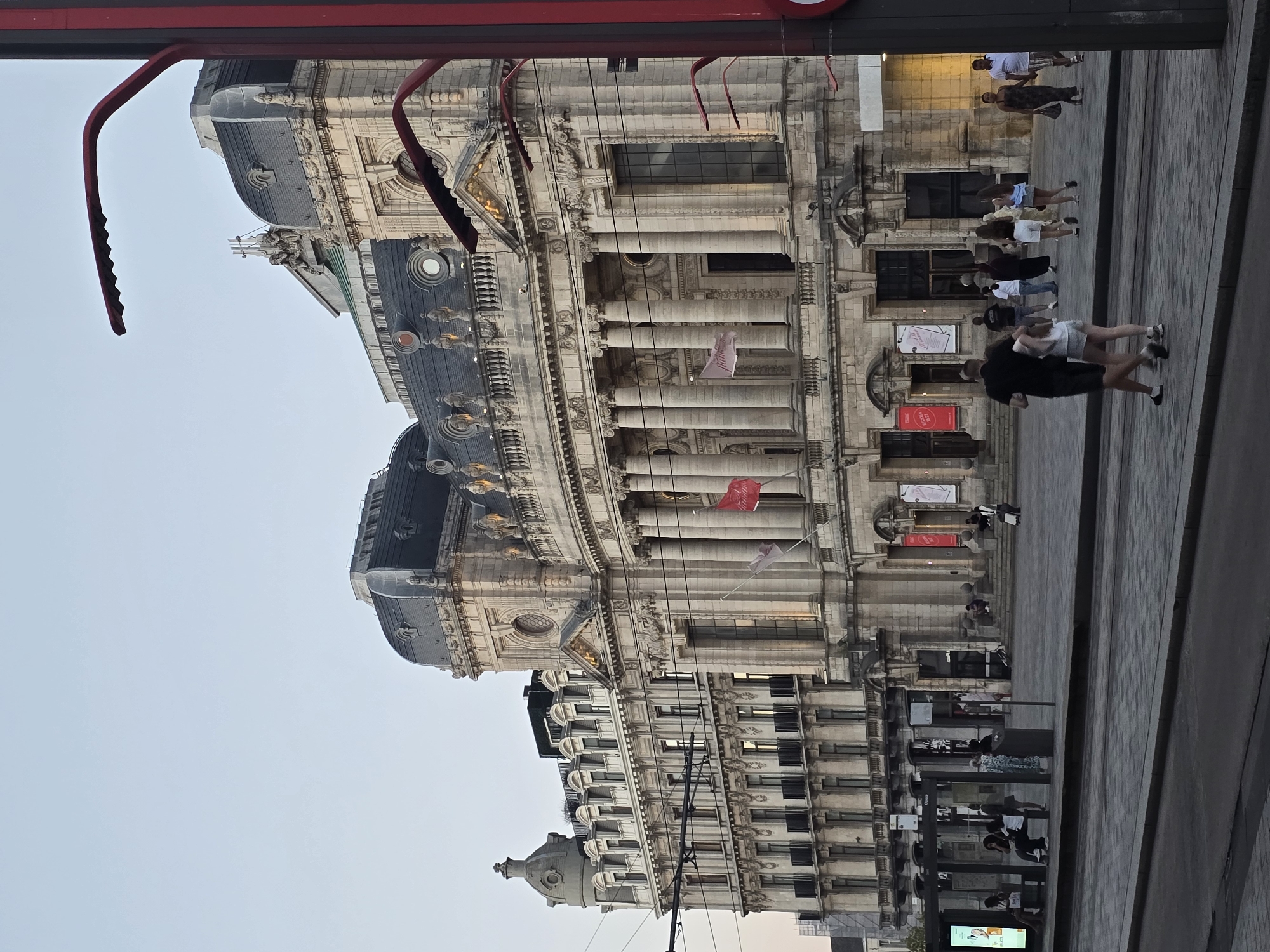 This is an outdoor, eye-level, full shot of the exterior of the Antwerp Opera House and a public plaza in front of it. The building's ornate stone facade is prominent, with multiple stories, arched windows, and columns. Red flags with white lettering are displayed. To the left, a tram stop is visible with a glass-enclosed shelter containing several people. The plaza has a paved surface and a few individuals are walking or sitting. A couple is walking towards the right of the frame in the foreground. A red lamppost is on the far right of the image. The sky is clear and a pale blue, suggesting it is either early morning or late evening.