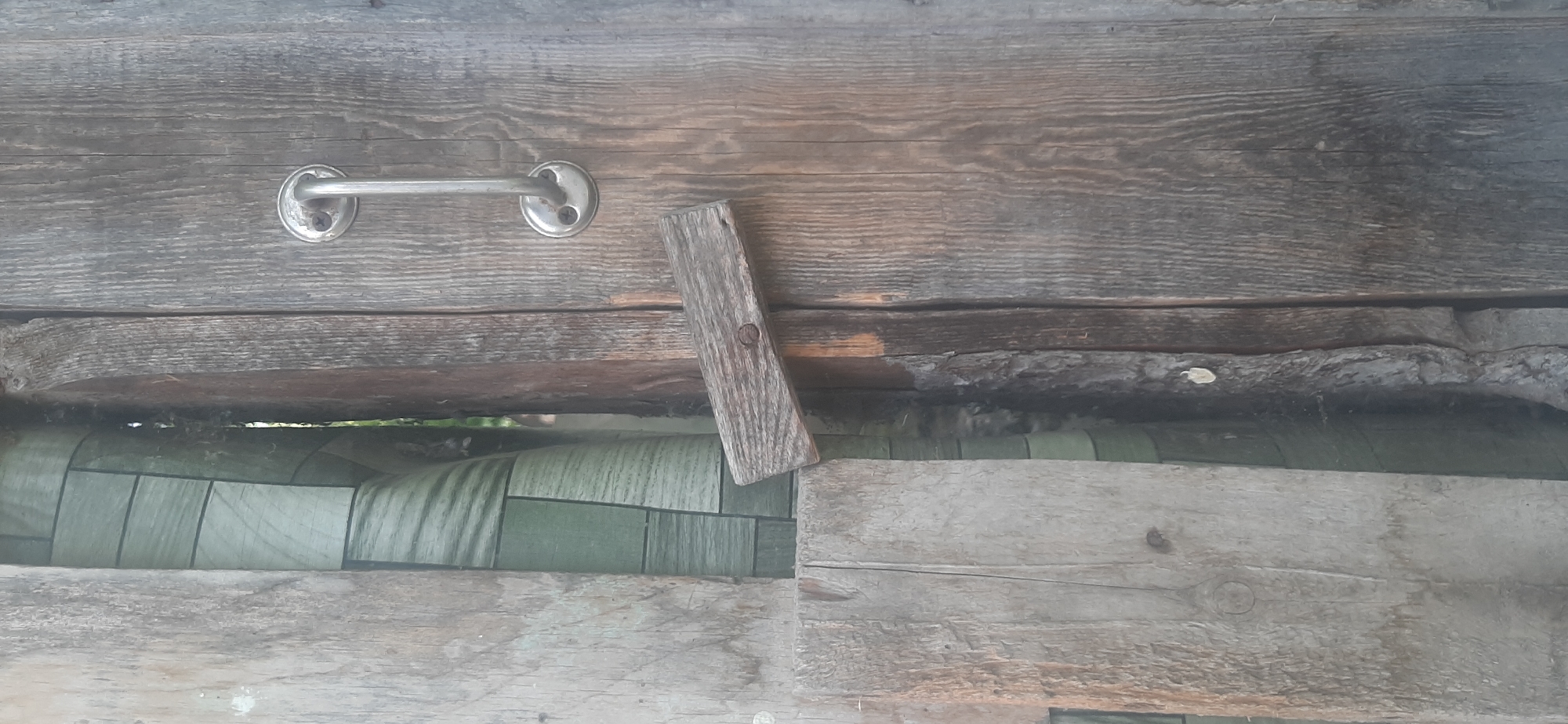 The image shows a close-up view of weathered wooden planks. A silver metal handle is attached to the upper plank with two visible screws. To the right of the handle, a small, rough-hewn piece of wood is affixed diagonally with a nail. Below the upper plank, a gap reveals a rolled-up material with a pattern of green rectangles and dark lines. This material appears to be some kind of matting or fabric. The lower part of the image is dominated by more wooden planks, some of which are wider and appear more worn than the upper ones. The overall impression is one of rustic, outdoor construction, possibly a shed, fence, or other utilitarian structure. The lighting suggests it is daytime, and there are no clear indicators of specific weather conditions. The scene is static, with no activity taking place. The location context provided is Yessentukskaya, Russia, which is consistent with the appearance of aged, natural materials. There is no visible text in the image.