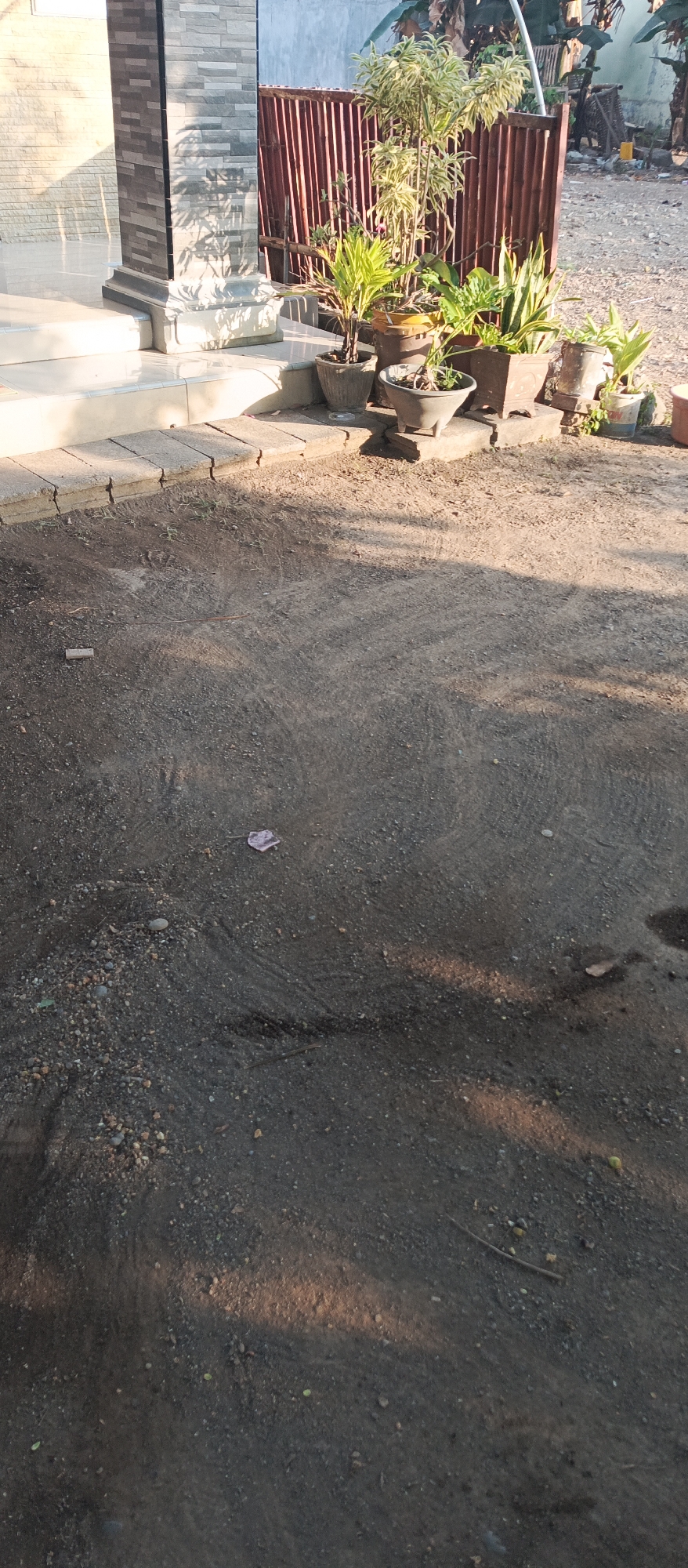 The image depicts an outdoor scene in Jatisari, Indonesia, focusing on the ground and a small cluster of potted plants near a building. The ground is composed of dark soil and small gravel, with tire tracks faintly visible in some areas. A small piece of pink debris lies on the ground in the mid-left portion of the frame. To the right, several potted plants of varying sizes are arranged on the ground and on small concrete blocks. One larger plant with variegated leaves stands prominently in a yellow pot. Behind the plants, a reddish-brown metal fence is visible. To the left, the base of a building with a textured stone-like facade and a set of steps leading up to it can be seen. Sunlight casts shadows across the scene, indicating it is likely daytime.