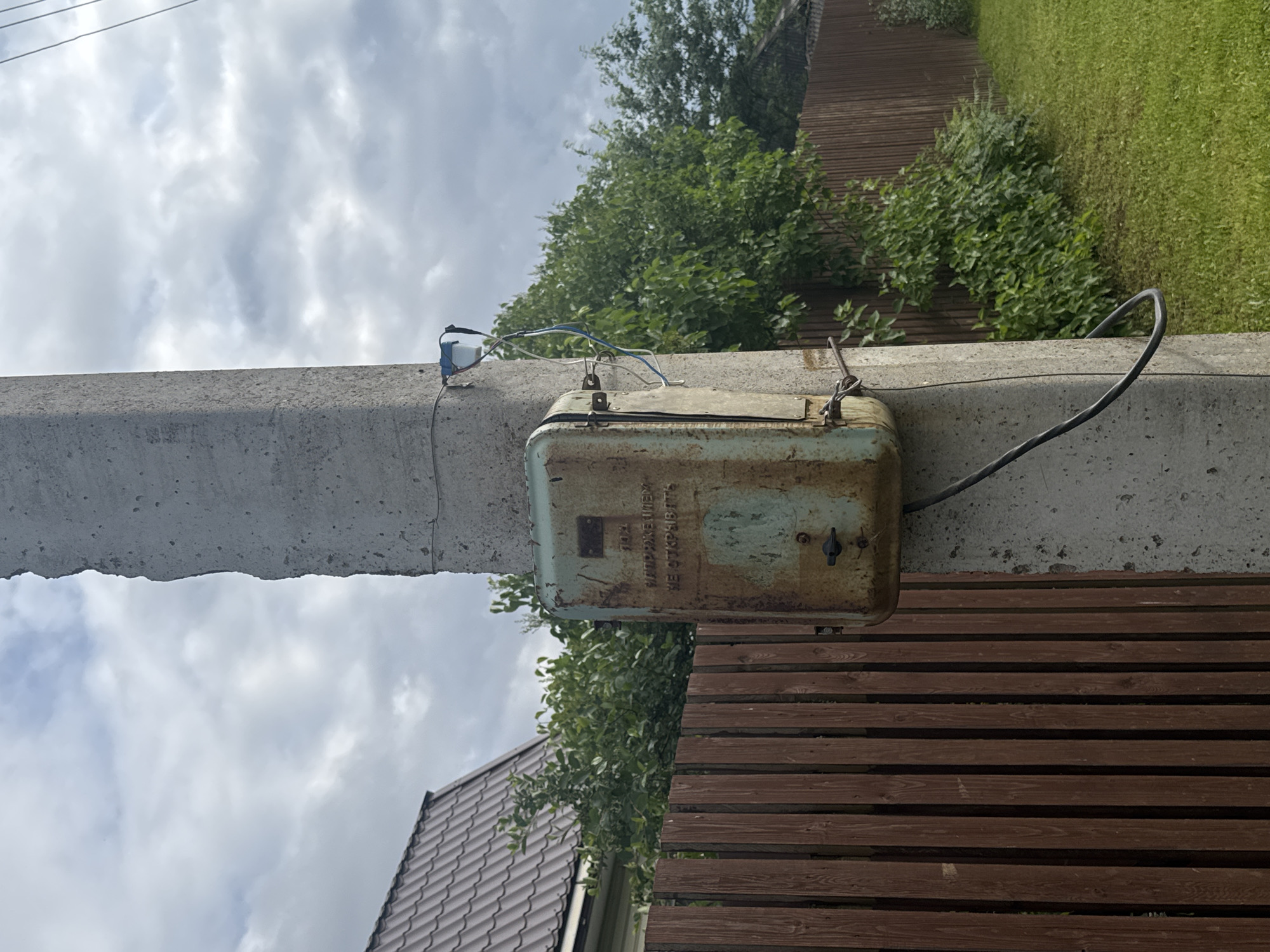 A weathered, rusted metal electrical box is attached to a concrete utility pole. The box has Cyrillic text on it which reads "702", "НАПРЯЖЕНИЕ ПОД", and "НЕ ОТКРЫВАТЬ". A black electrical cable descends from the box and is secured to the pole. A small blue and white object with wires is attached higher up on the pole. In the background, a brown wooden fence runs along the side of the property, with lush green trees and bushes behind it. A portion of a house with a brown shingled roof is visible to the left. The ground is covered in green grass. The sky is overcast with white and gray clouds. The scene appears to be in a rural or suburban setting, possibly in the village of Borodulino, Russia, given the Cyrillic text. There are no people visible in the image.