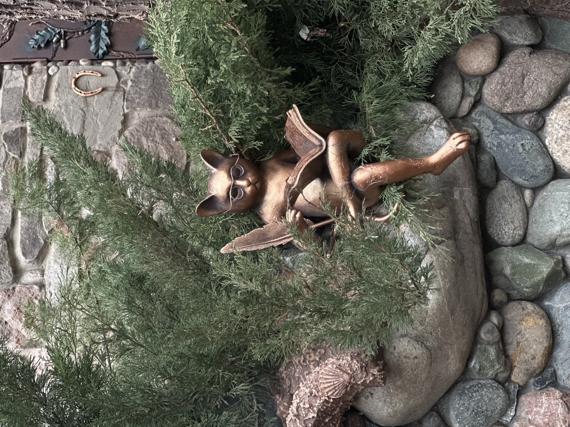 The image shows a bronze statue of a cat wearing glasses and reading a book, nestled among green bushes and gray rocks. A horseshoe is visible on a stone wall in the background. The image does not provide information about the location of Izhevsk, Russia.
