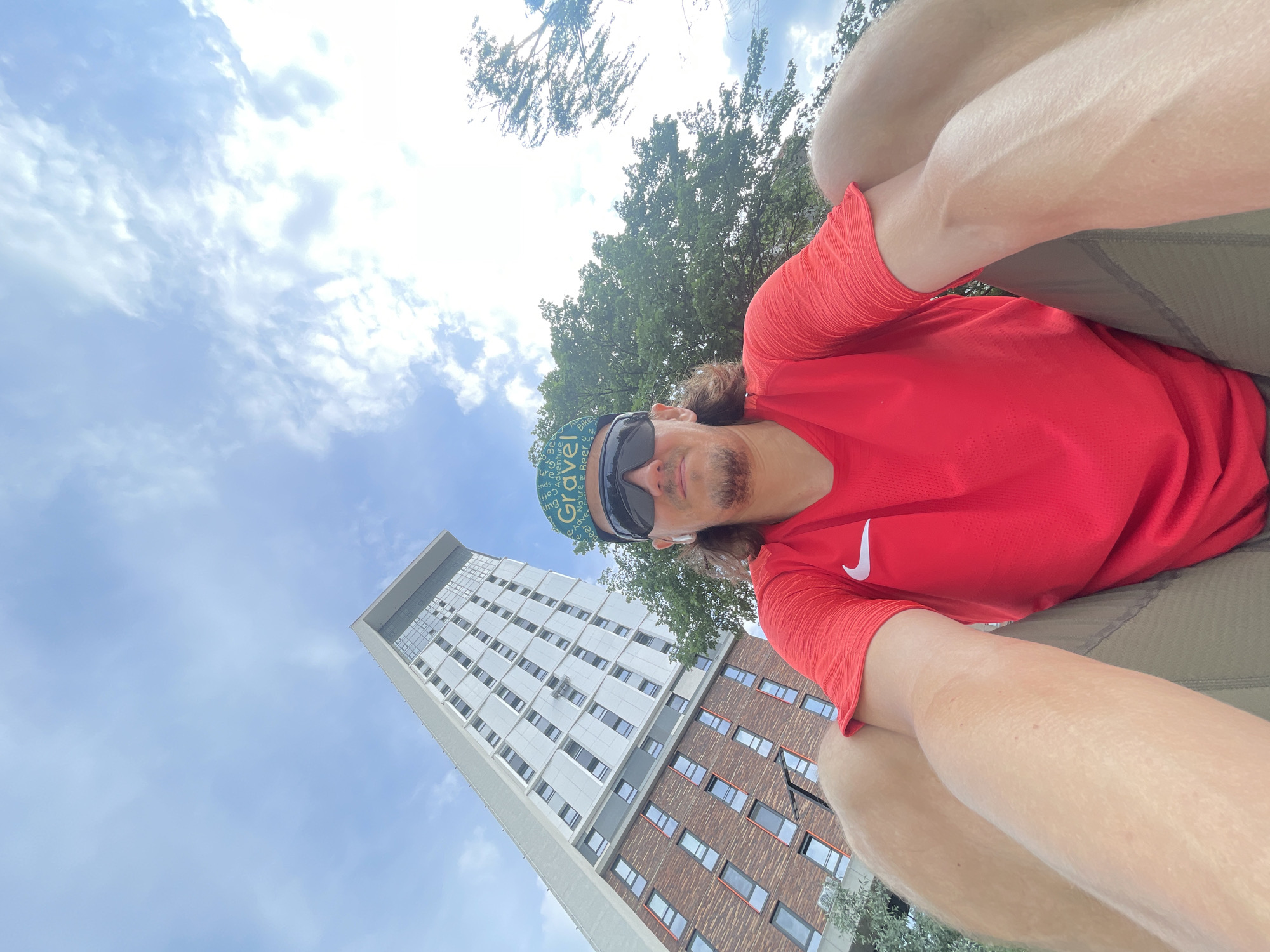 A low-angle, outdoor selfie captures a man from a perspective looking upwards. He is an adult male with medium-length brown hair and stubble, wearing dark sunglasses, a bright red athletic shirt with a white Nike swoosh on the left chest, and dark shorts. He also wears a light blue cap with the word "Gravel" prominently displayed, along with other partially visible words like "Nature," "Bike," and "Adventure." His arms and legs are visible, suggesting he is in a seated or squatting position.
The background is dominated by a clear blue sky with scattered white clouds, indicating daytime with bright sunshine. To the left, a tall, modern building rises into the sky, featuring a multi-story facade composed of light-colored and red-brown sections with numerous rectangular windows. Green trees with dense foliage are visible in the mid-ground and background, particularly behind and to the right of the man. The scene suggests an urban or semi-urban outdoor setting, possibly a park or campus area next to architectural structures.