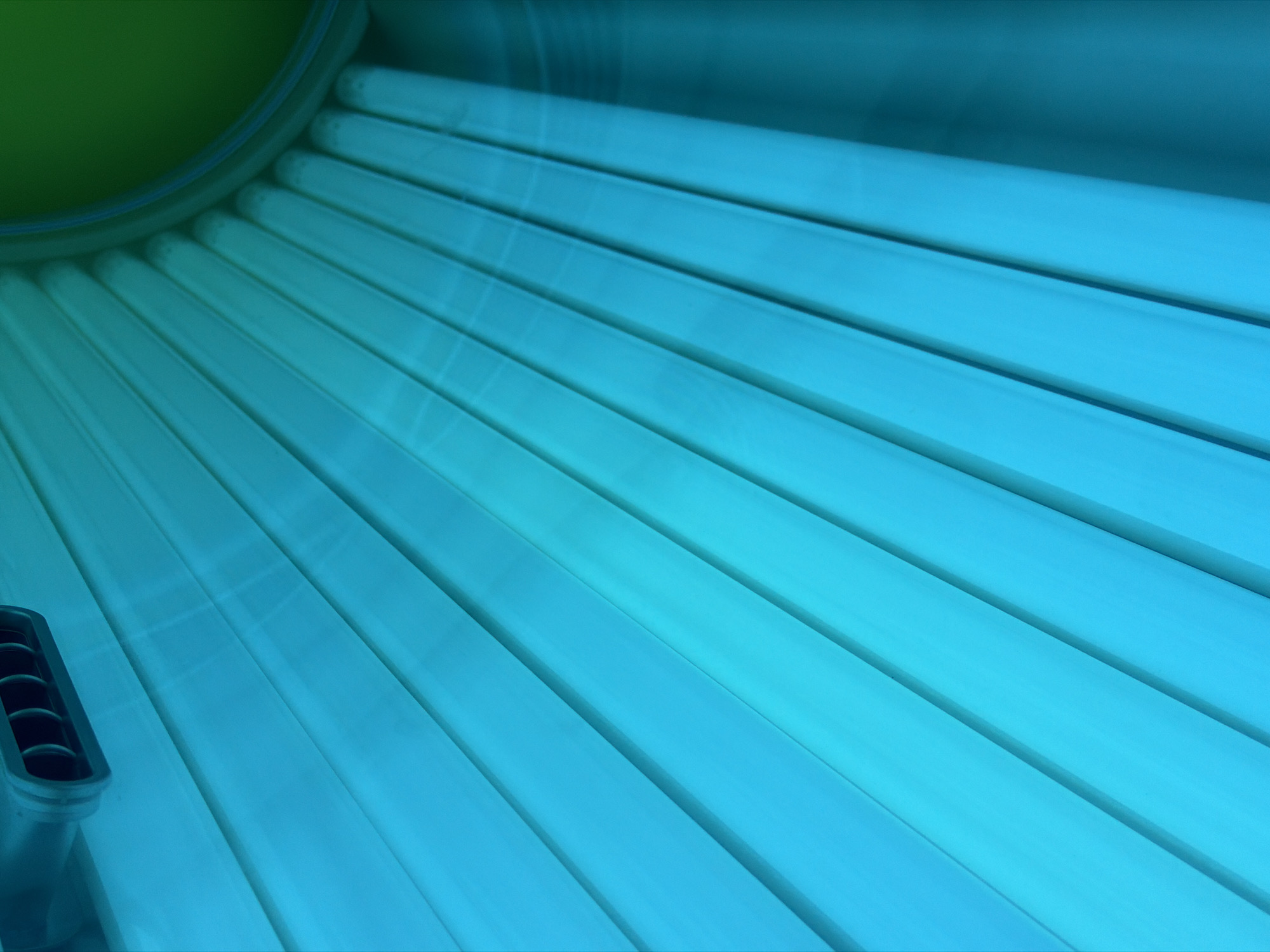 The image shows the inside of a tanning bed. The bed is illuminated by bright blue fluorescent lights arranged in a fan-like pattern. The bottom right corner of the image reveals a curved, green surface, likely the bed's interior. A grey vent is visible in the upper right corner. The scene is abstract and does not provide any information about the city of Kyiv, Ukraine.