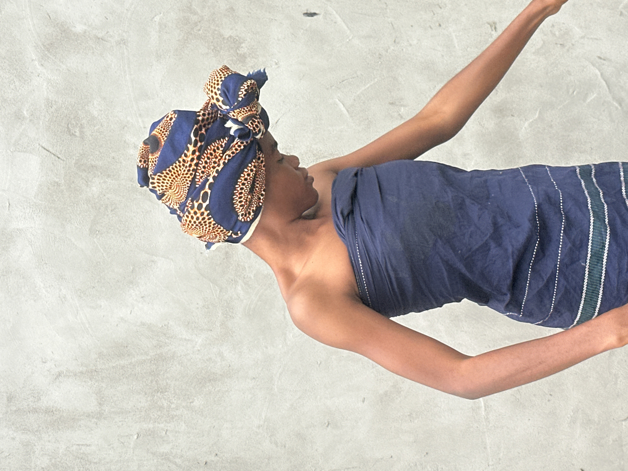 A young Black female stands in front of a textured, light gray concrete wall. She is wearing a dark blue, strapless garment wrapped around her chest, with horizontal stripes of white and green embroidery near the bottom. A patterned headwrap, featuring navy blue, orange, and beige circular designs, is tied around her head and neck. Her head is bowed, and her gaze is directed downward. Her right arm extends outwards to the right. The lighting suggests an outdoor setting, likely during the day, with soft, diffused light casting minimal shadows. The background is a plain concrete wall, indicating an urban or semi-urban environment. This image was captured in Badagry, Nigeria.