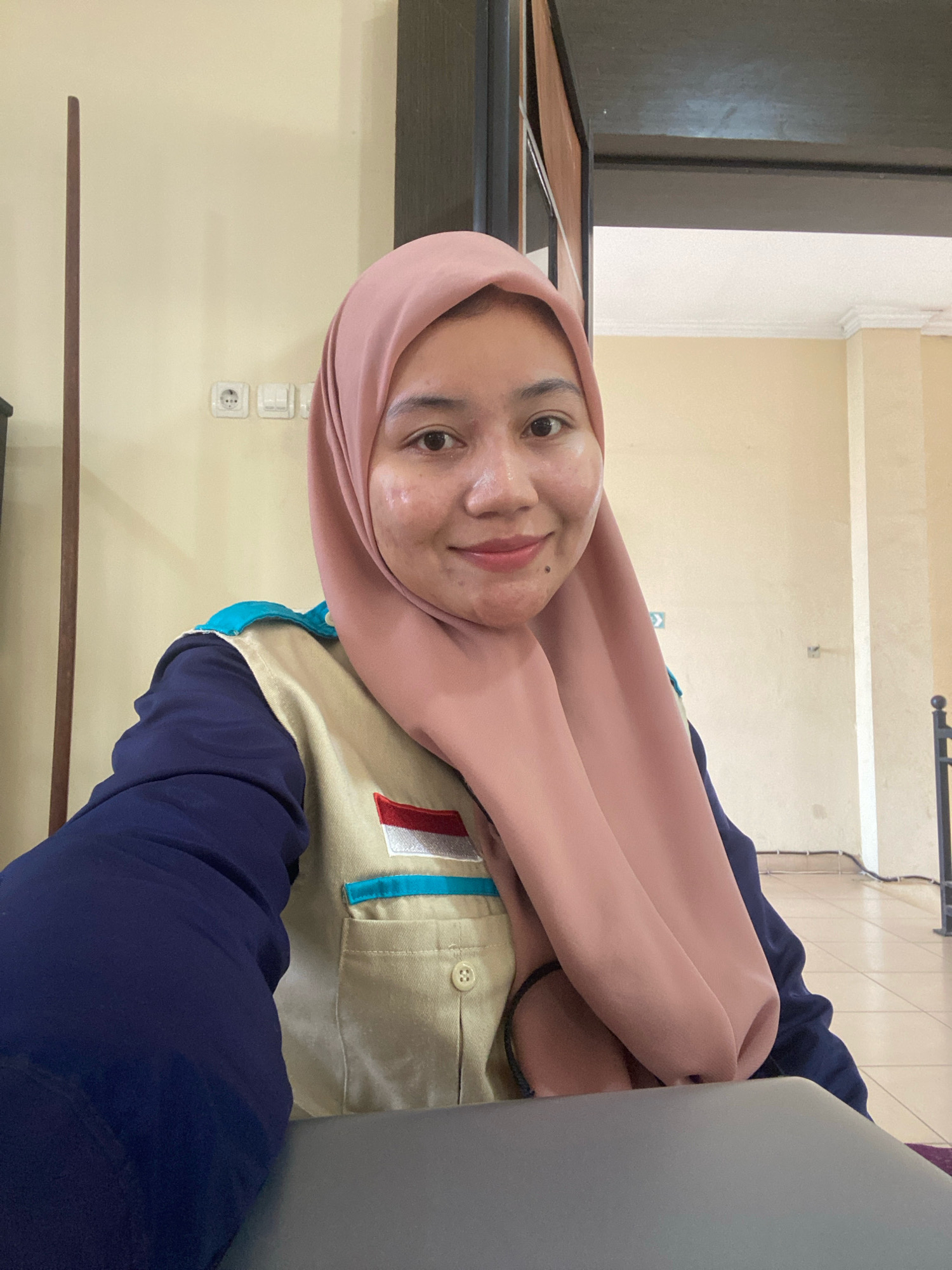 A young woman wearing a pink hijab and a tan vest over a navy blue long-sleeved shirt is looking at the camera, smiling. The vest has a patch on the left side that appears to be the Indonesian flag. She is sitting in what seems to be an office or classroom setting, with a light yellow wall behind her and a dark wooden partition to her right. Electrical outlets are visible on the wall. In the foreground, the edge of a gray laptop is visible. The overall lighting suggests an indoor environment, possibly during the daytime. The location is identified as Banjarbaru, Indonesia.