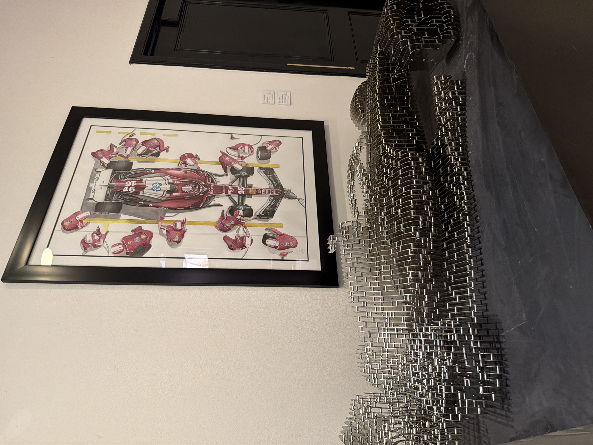 A framed print of a race car in a pit stop is displayed on a white wall, positioned below a dark framed object. To the right of the print, a sculpture consisting of metal grids is partially visible. The location is San Bernardino Tlaxcalancingo, Mexico, although this cannot be verified from the image alone.