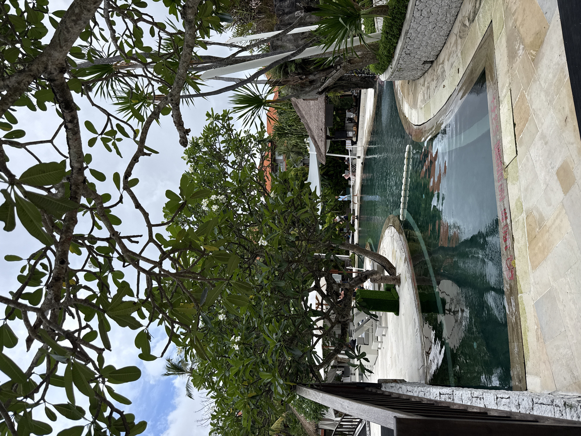 The image depicts a swimming pool area in Nusa Dua, Indonesia. The pool is surrounded by white paving stones and includes a curved section. A line of buoys runs across the pool. Trees with green leaves frame the scene, partially obscuring the sky, which shows some blue patches. A building with a thatched roof and white awnings is visible in the background, with some chairs and tables underneath. People are present near the building and along the pool. A wooden railing is visible in the lower foreground.