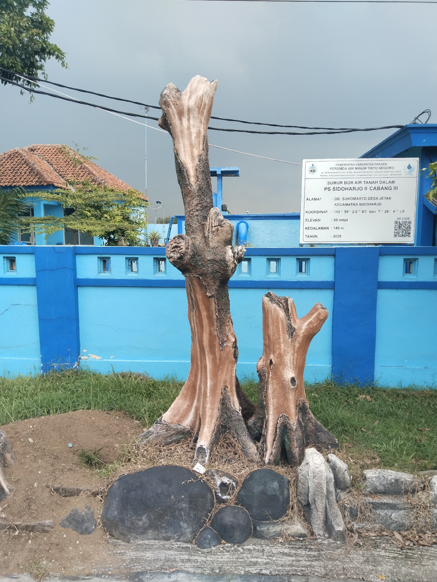 The image captures a scene in Sidoharjo, Indonesia, showcasing a display of two sculpted tree trunks positioned in front of a blue wall. Behind the wall, a building with an orange tile roof is partially visible. To the right of the tree trunks, a white sign provides information about the "Sumur Bor Air Tanah Dalam PS Sidoharjo II Cabang III," indicating this is a groundwater well site. The sign also includes the address, coordinates (110° 59' 2.8" BT and 7° 26' 4" LS), elevation (80 mdpl), depth (140 m), and the year (2020). The base of the tree trunks is surrounded by black and grey rocks, set on a concrete surface, with green grass visible on the ground behind the rocks and in front of the blue wall. Overhead, electrical wires stretch across a cloudy sky.