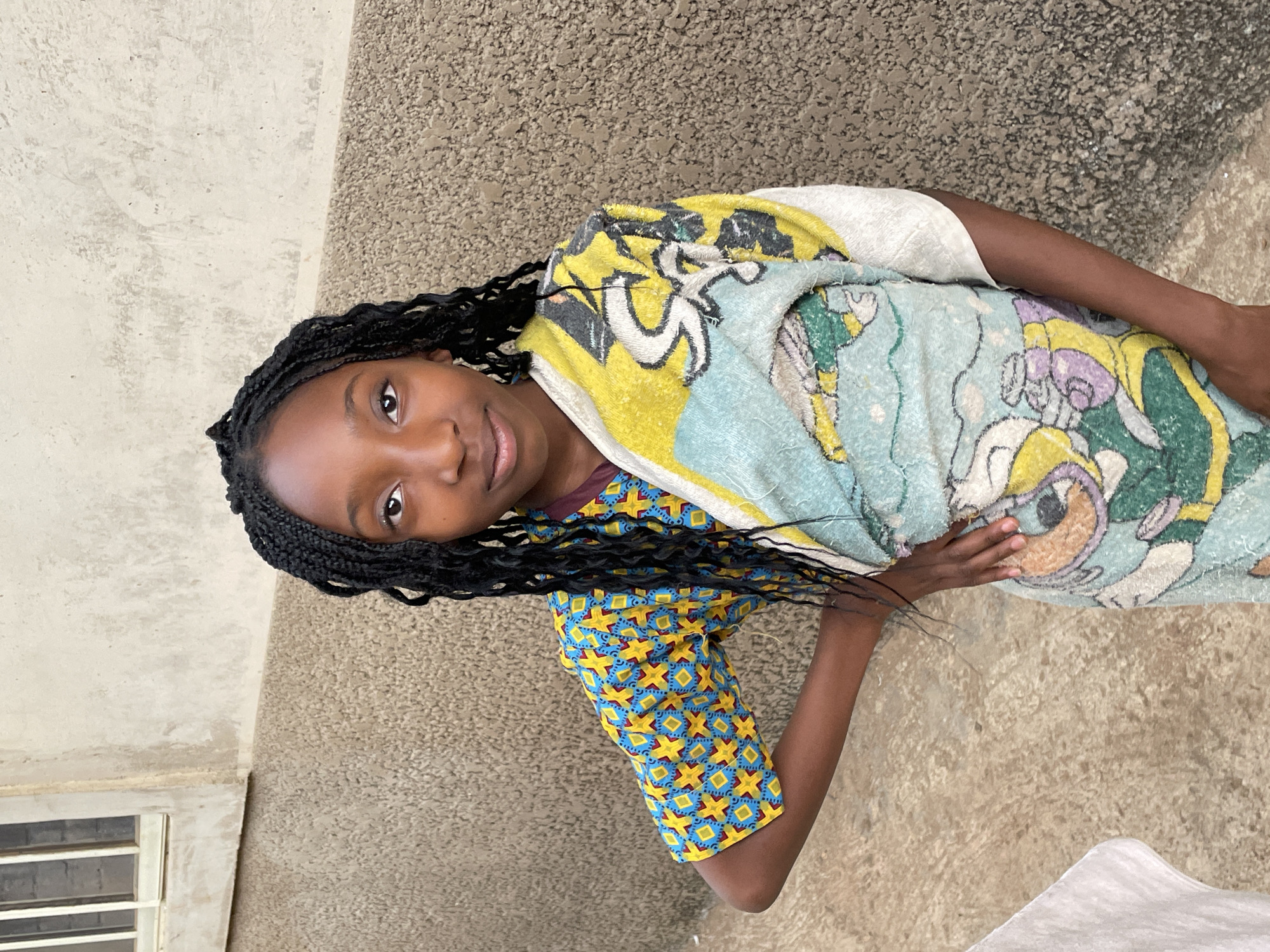 A young Black girl with braids is standing outside in Jos, Nigeria. She is wearing a blue and yellow patterned shirt and a light blue and yellow cartoon-print towel draped around her shoulders. Her right hand is resting on the towel. The background consists of textured concrete walls and a window with white frames. The ground appears to be concrete as well.