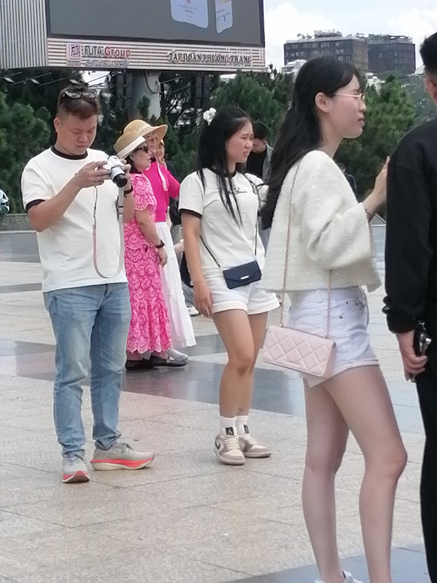 In Da Lat, Vietnam, on a paved walkway, several individuals are captured. In the foreground, a man in a white t-shirt and blue jeans holds a camera. Beside him, a woman in a pink, ornate dress and a straw hat stands. To the right, a woman in a white top and white shorts carries a light pink quilted purse. Further to the right, another person in black clothing is partially visible. The background features lush green trees and buildings. A large billboard with Vietnamese text is mounted above the scene.