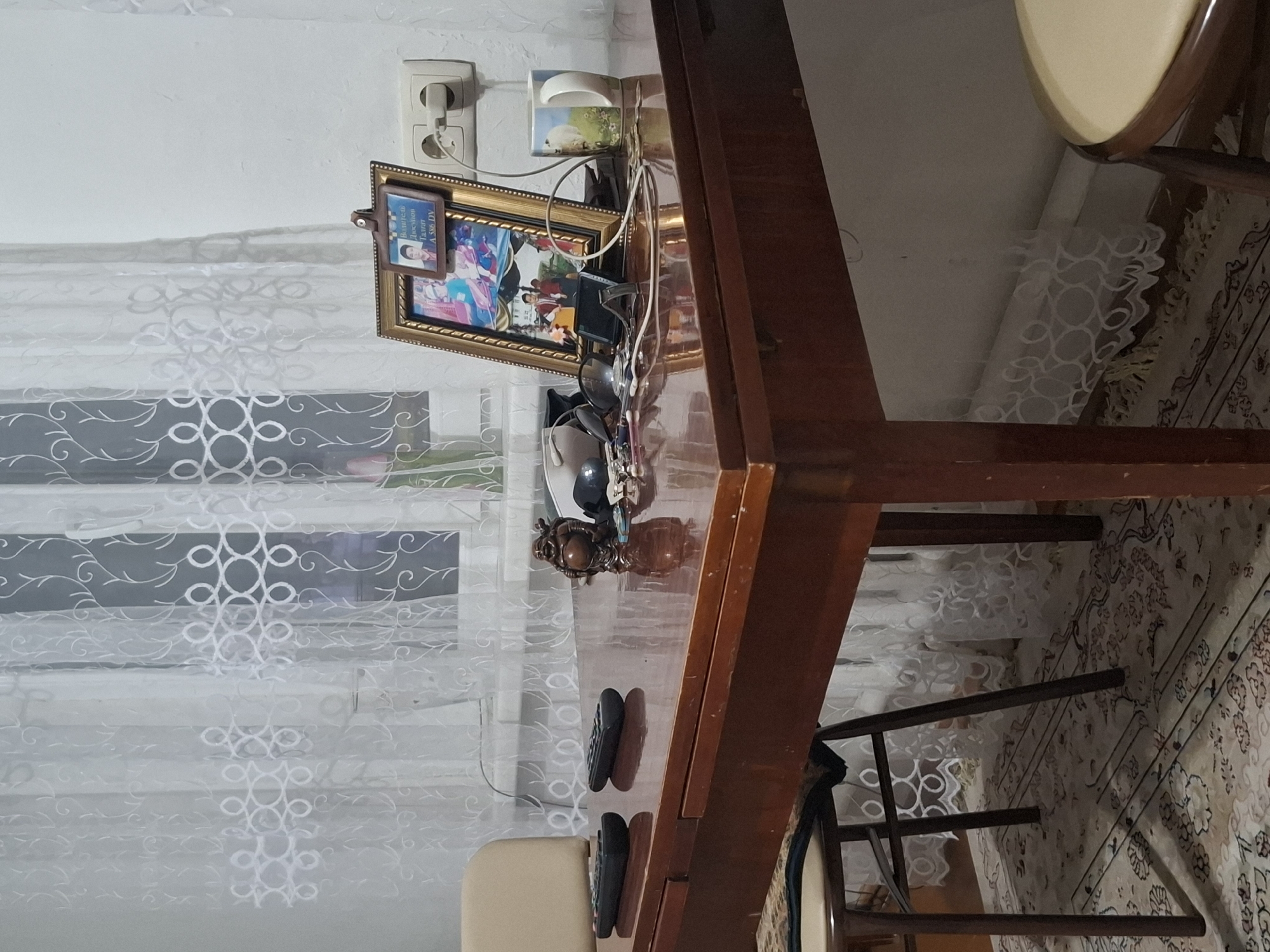A dimly lit, indoor scene depicts a wooden table with several objects on its surface. Two black remote controls are placed on the left side of the table. To the right of the remotes, a small, dark brown statue of a laughing Buddha is positioned. Further to the right, a framed photograph stands upright, displaying three images within it. Adjacent to the photograph is a white mug with a pastoral scene. Cables and a pair of sunglasses are also visible on the table. Behind the table, a white sheer curtain with a patterned design covers a window. The table itself appears to be made of polished wood, with its surface reflecting the ambient light. The floor beneath the table is covered by a patterned rug. The location is identified as Almaty, Kazakhstan.
