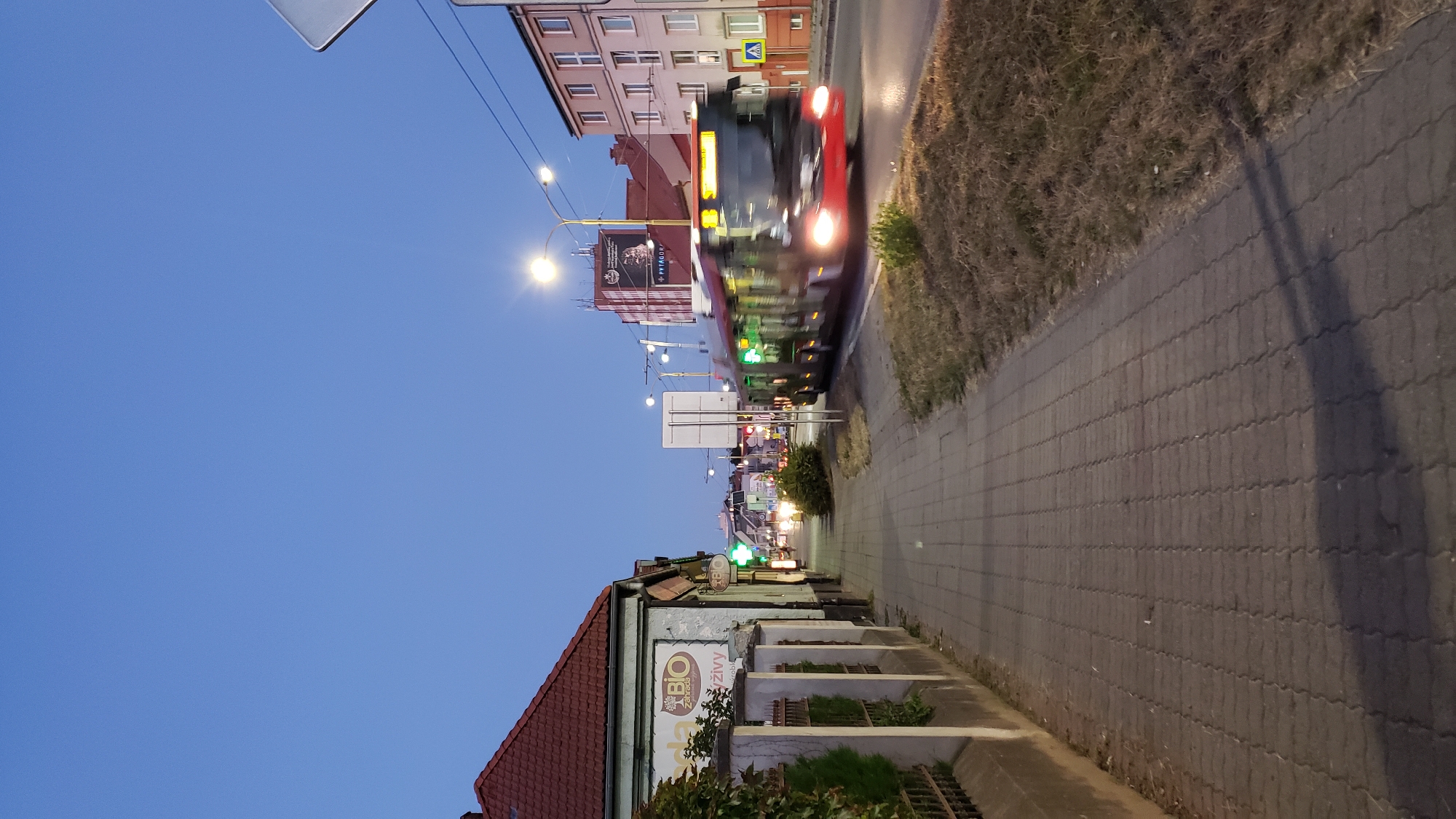 This is a vertical, eye-level shot of a street scene in Prešov, Slovakia. The image captures a sidewalk on the left, with a building facade and a fence. On the right, a street with a moving bus and buildings. The sky is a deep blue, indicating dusk or early evening.
On the left side, a light-colored building with a red-tiled roof is visible, featuring a sign with "BIO" and "Záhrada" (garden in Slovak). The fence in front of the building is made of concrete posts and metal bars. A patch of green foliage grows along the fence. The sidewalk is paved with interlocking gray bricks.
The right side of the image shows a street with a modern articulated bus in motion, its headlights on. The bus has a red and dark gray exterior and its windows are illuminated. The side of the bus displays green illuminated signs. The street is flanked by dry, grassy verges. In the background, there are multi-story buildings and streetlights. One building has a large billboard with text including "+PYTAGOR". A yellow pedestrian crossing sign is visible near the right edge of the frame.
The overall atmosphere is urban, with the fading light of dusk creating a sense of transition. The presence of the bus suggests public transportation is active. The scene is devoid of any identifiable individuals, but the focus is on the urban infrastructure and the movement of the bus.
