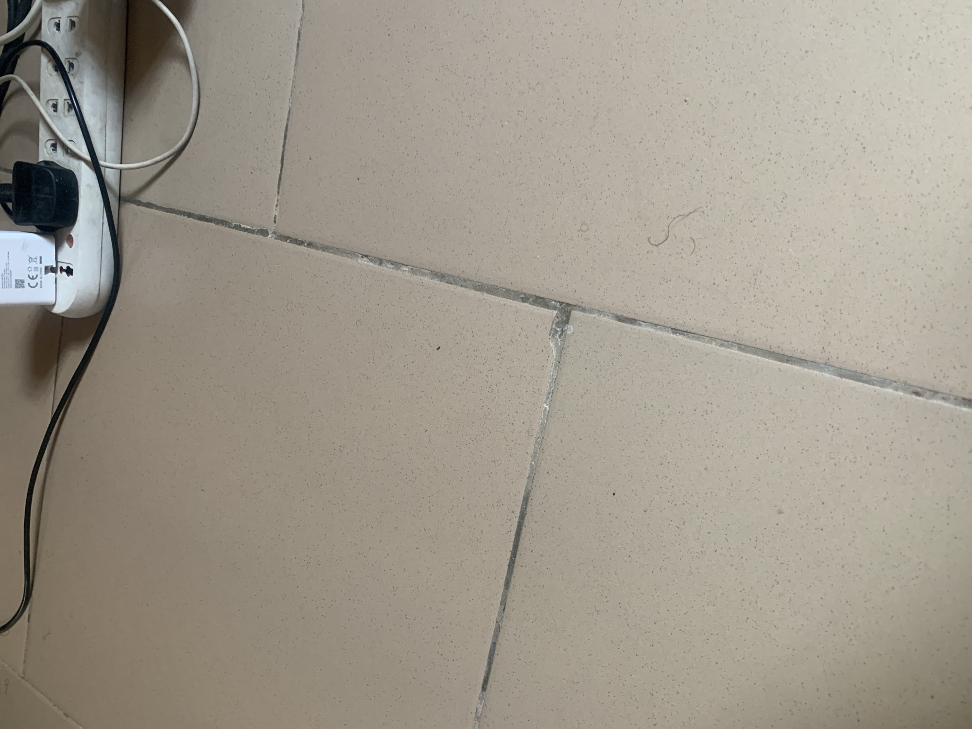 The image displays a close-up view of a tiled floor with visible grout lines. In the upper portion of the image, a white power strip with several electrical outlets is present, along with various power adapters and cables plugged in. One adapter is a white USB charger with "CE" markings. The floor tiles are a neutral, light beige color with small speckles. A single strand of dark hair is visible on the tiles in the lower right quadrant of the image. The scene appears to be indoors, likely in a residential or commercial setting. No specific location within Jauro Yinu, Nigeria can be identified from the image itself.