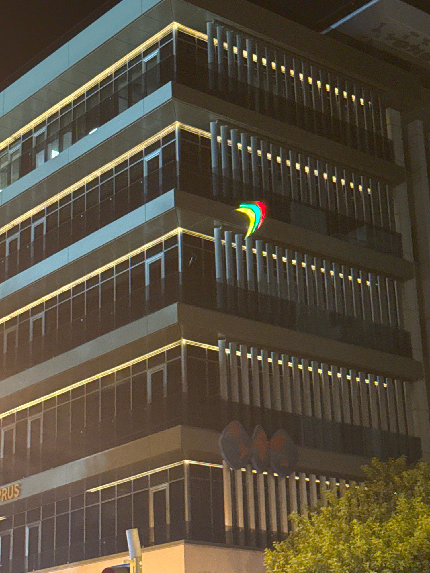 The image shows a modern, multi-story building at night, illuminated by external lighting. The building has a facade with numerous windows and vertical louvers. A colorful, abstract logo, appearing as a series of curved shapes in red, green, and blue, is mounted on the building's exterior, likely a sign. Below this logo, on a lower level of the building, are three circular objects, possibly traffic signals or artistic elements, in shades of blue and orange. The lower part of the building displays the partial text "CYPRUS" in orange lettering. A lush green tree is visible on the right side of the image, with its foliage partially obscuring the lower floors of the building. The overall scene suggests an urban environment in Limassol, Cyprus, likely in a commercial or office district.