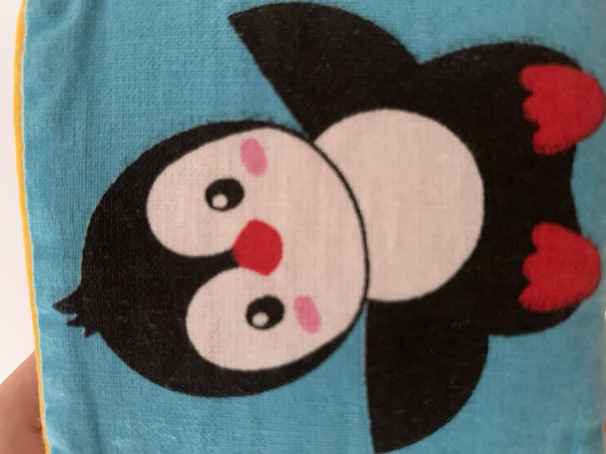 A close-up shot of a cartoon penguin on a light blue fabric surface. The penguin has a white belly and face, black wings, head, and feet, and a red beak and cheeks. The fabric has a yellow edge at the top. The image is a photograph and is not located in Stavropol, Russia.
