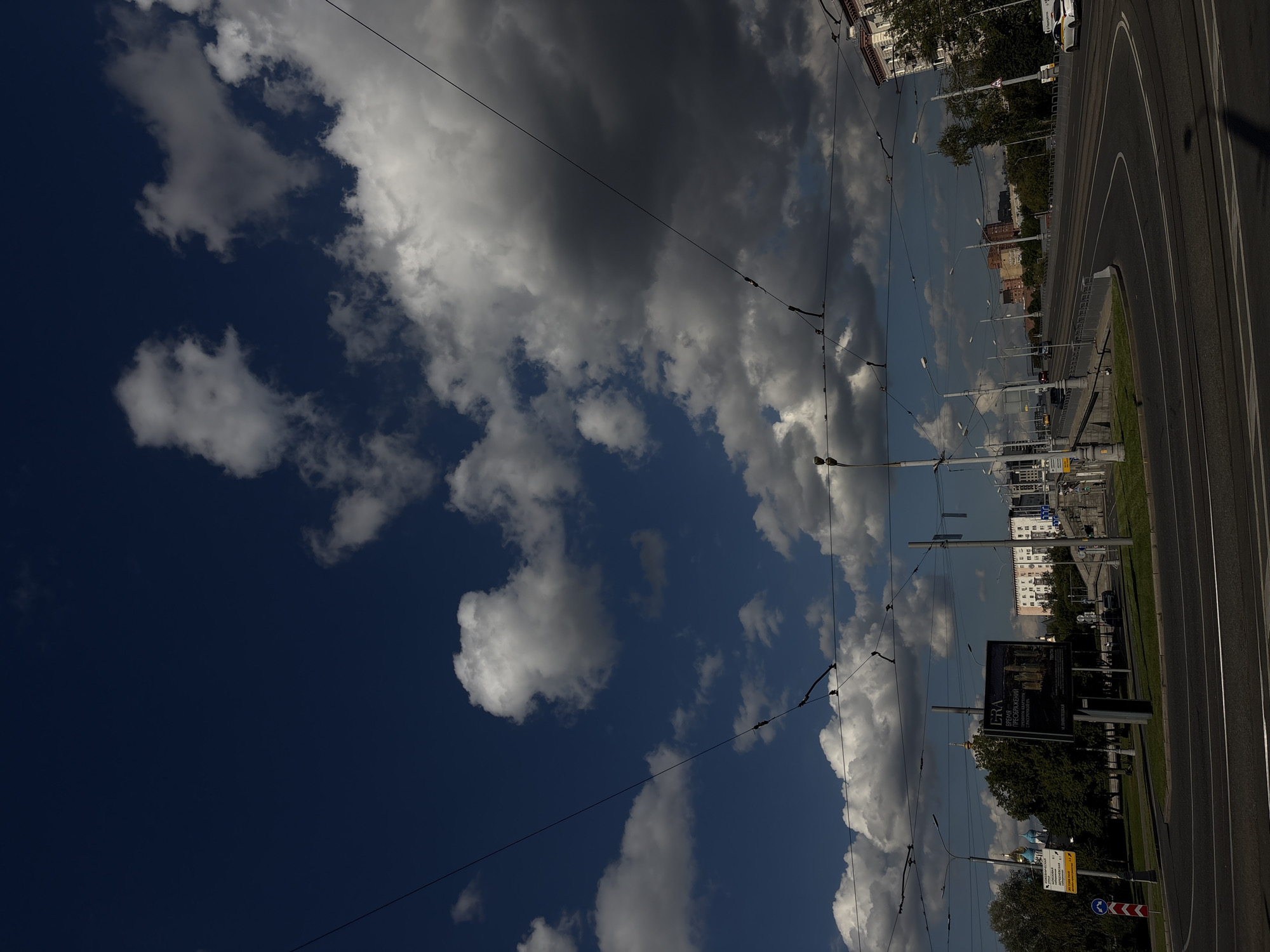This is a photograph of a road with tram lines in Moscow, Russia, under a cloudy sky. In the foreground, there is a wide asphalt road with visible tram tracks. On the left side of the road, there is a green park area with trees and a billboard advertising "ERA" housing. Beyond the park, blue-domed buildings and more trees are visible. The road curves to the right, leading towards a bridge or elevated section with several lanes of traffic. Cars, including a taxi, are seen on the road. Overhead, tram wires stretch across the scene, supported by poles. The sky is a deep blue, with dramatic, large white and grey clouds casting shadows. The overall impression is of a city environment on a partly cloudy day, with a focus on urban infrastructure and the sky.