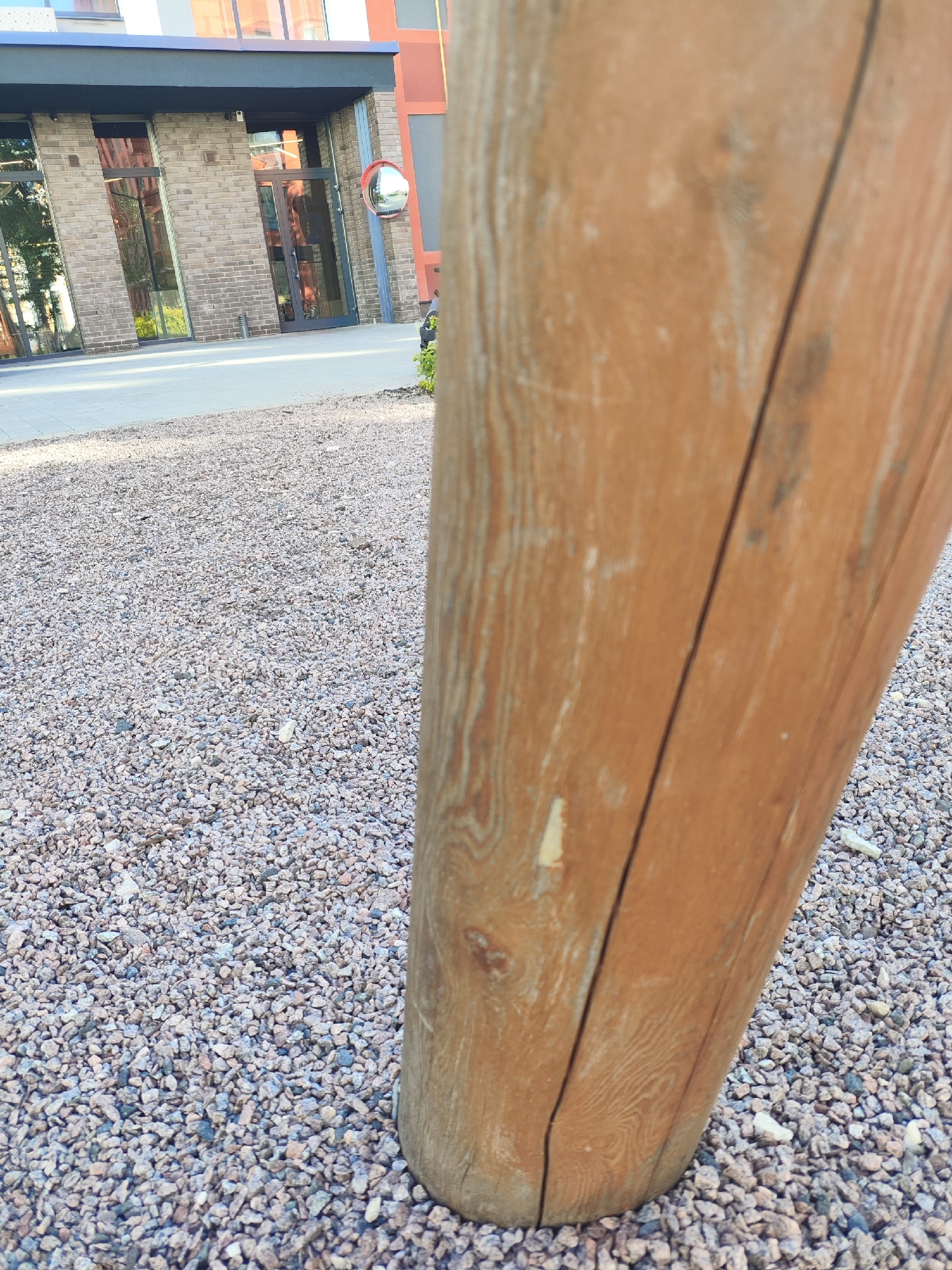 The image shows an outdoor scene with a large wooden post in the foreground, taking up the right side of the frame. The post is a light brown color with visible wood grain and a vertical split. The ground is covered in small, multi-colored gravel. In the background, there is a modern brick building with large windows and dark frames. Some greenery is visible near the building. A convex mirror is mounted on the side of the building near one of the entrances, reflecting a small portion of the surroundings. The scene appears to be an exterior walkway or courtyard. The lighting suggests it is daytime, with shadows cast on the ground. There is no text visible in the image. The overall impression is one of an urban or commercial outdoor space.